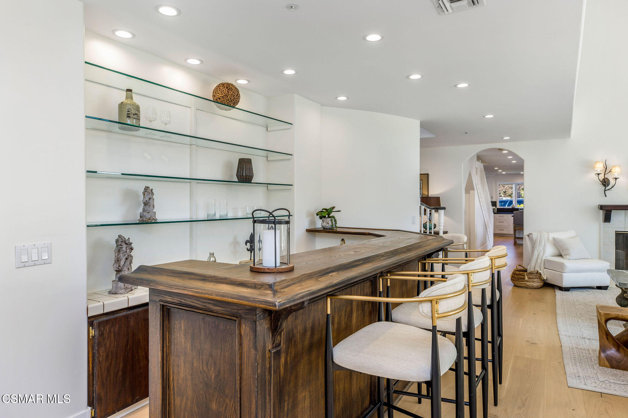 Dark wood bar with upholstered stools, glass display shelves, and an arched opening to the living area.