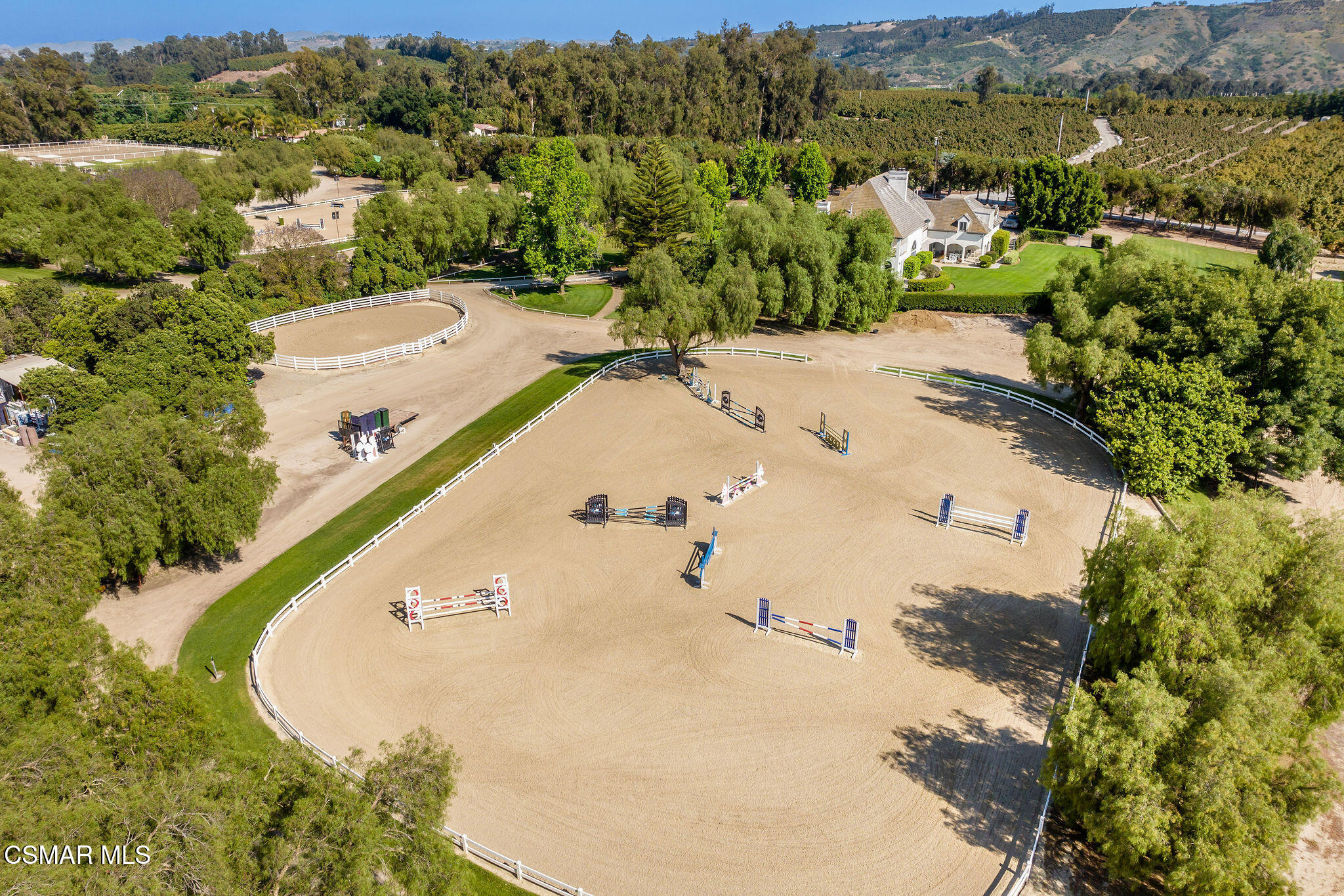 Aerial view of an equestrian estate with a large jumping arena, white-fenced rings, and a house.