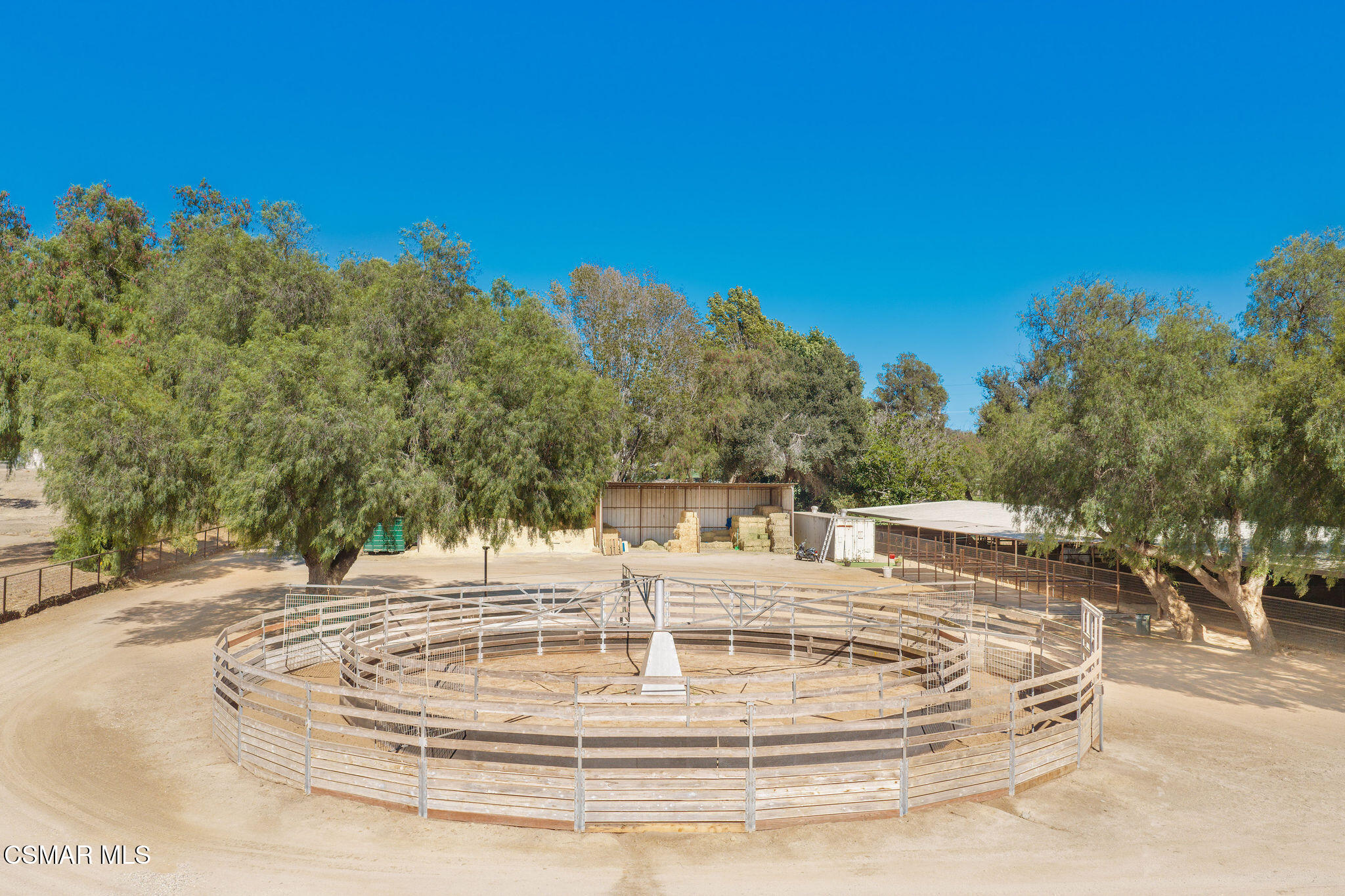 Circular wooden horse pen in front of hay storage and covered enclosures beneath mature trees.