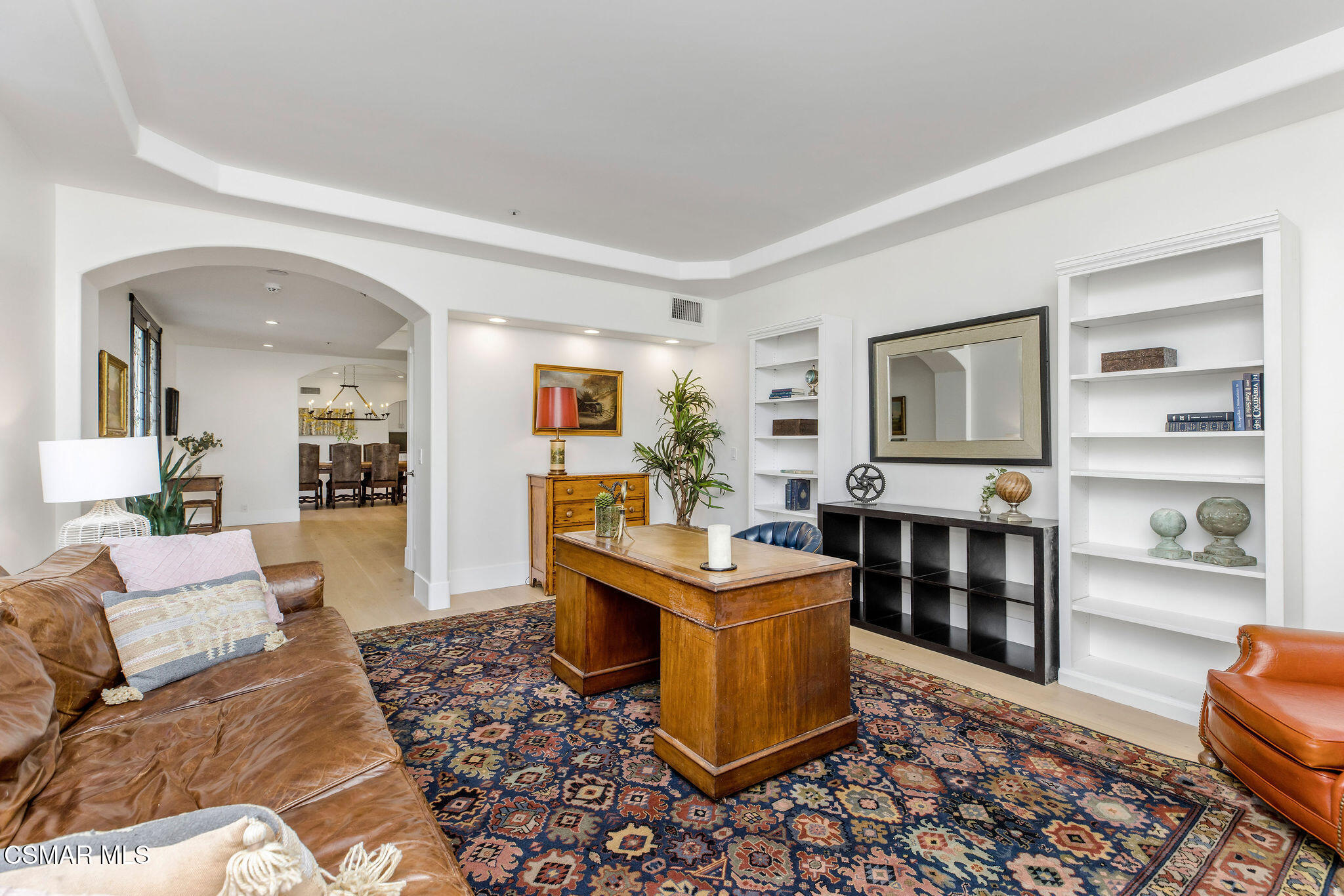 Furnished home office with a large wooden desk, built-in shelves, leather sofa, and patterned rug.