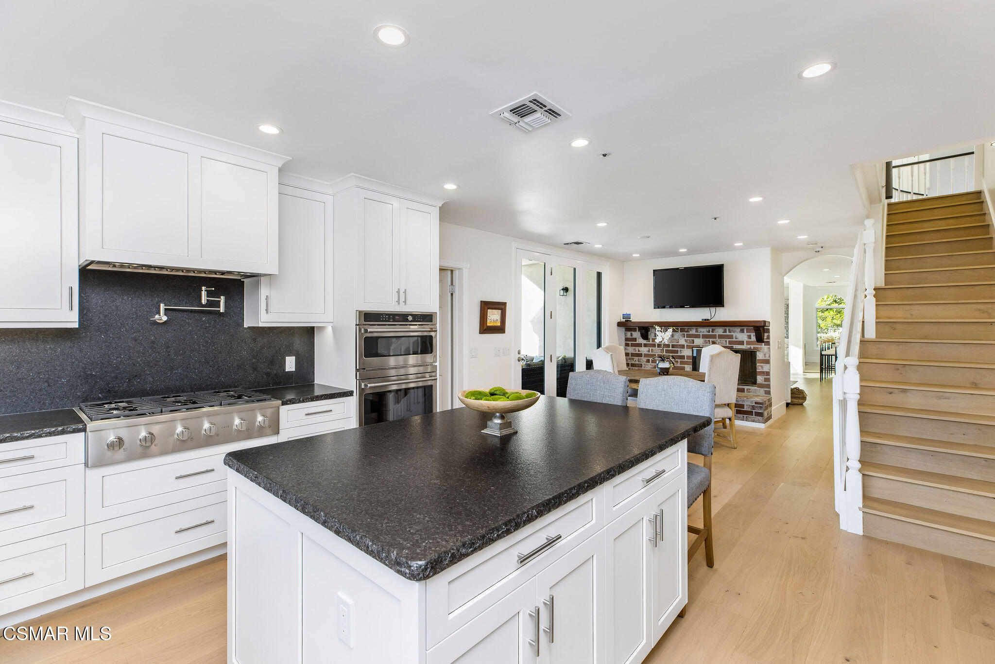 Open-plan kitchen with a large dark stone island, white cabinetry, brick fireplace, and wood staircase.