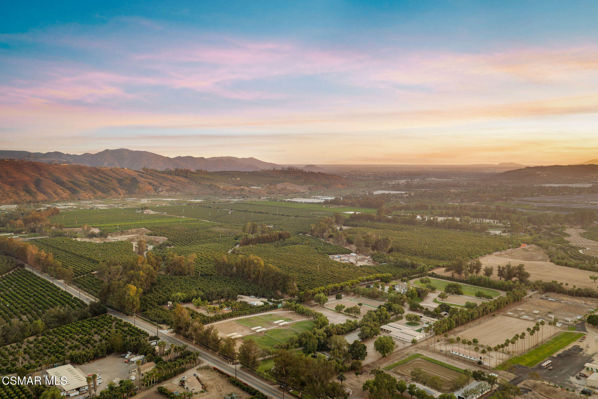 Aerial view of orchards and fields across a valley, with sports courts and mountains beneath a sunset sky.