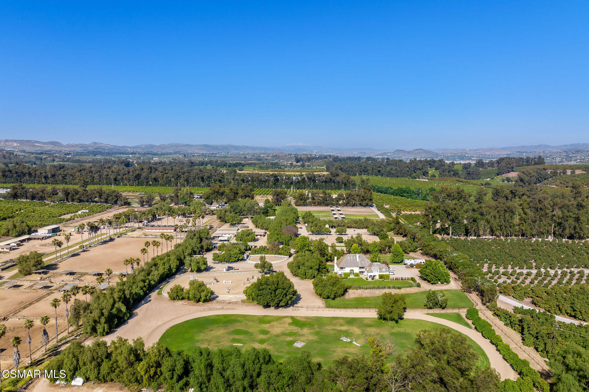 Aerial view of a country estate with a white house, riding ring, paddocks, and surrounding orchards.