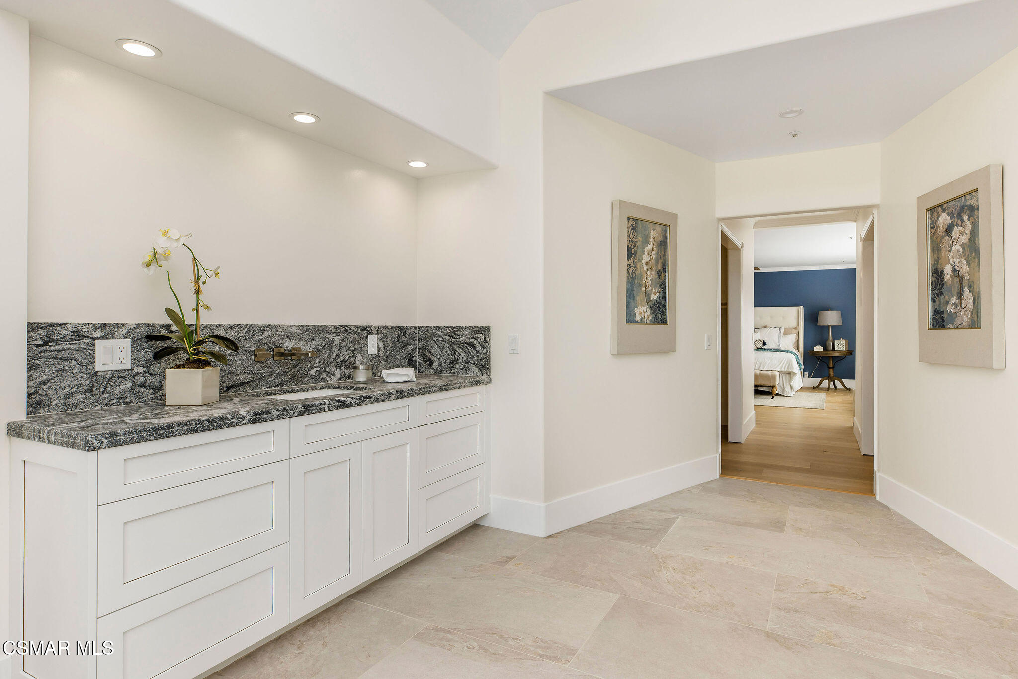 Bathroom vanity with a gray stone counter and orchid, looking through a doorway into a blue-accented bedroom.