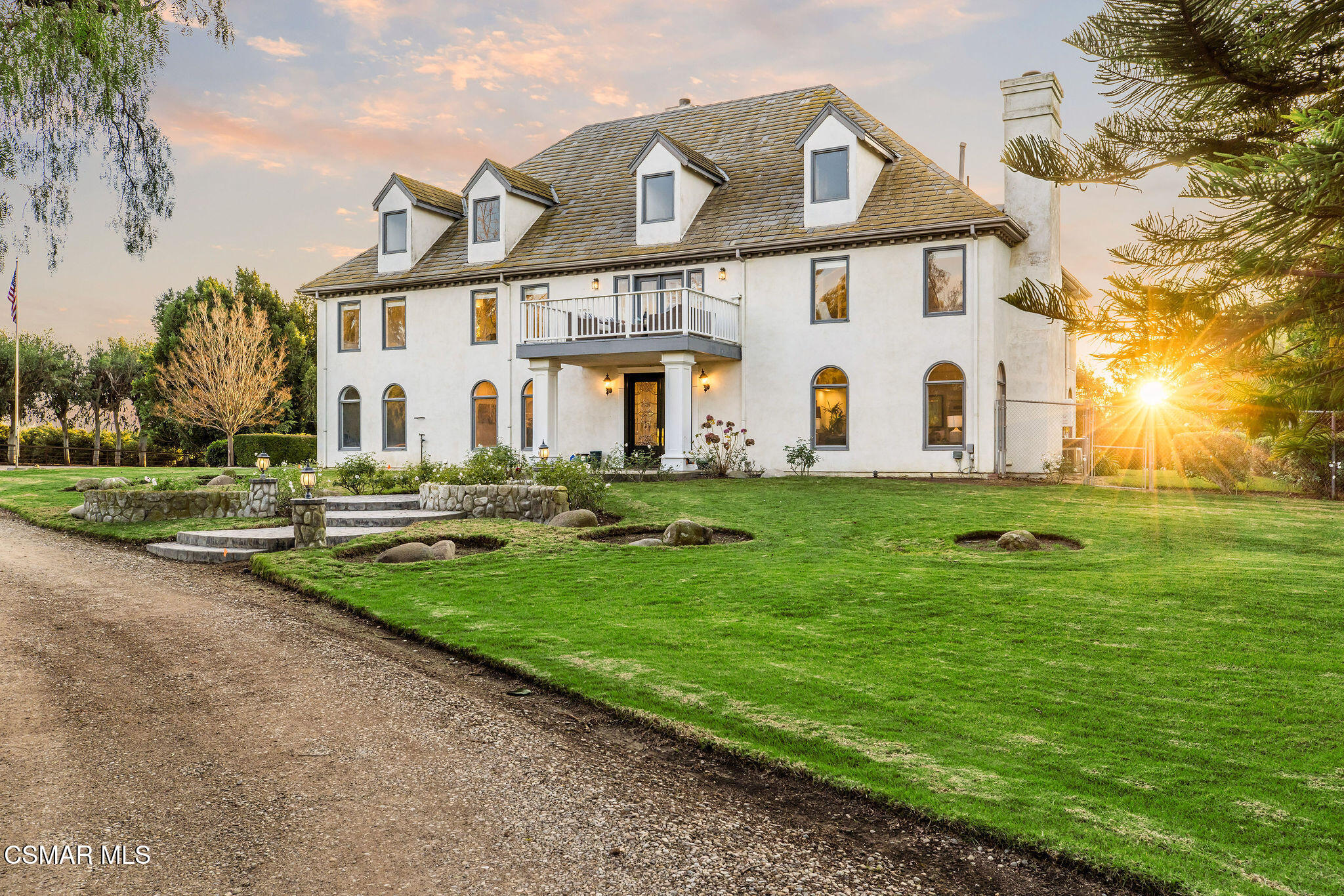 White two-story house with dormer windows and balcony, framed by a green lawn at sunset.