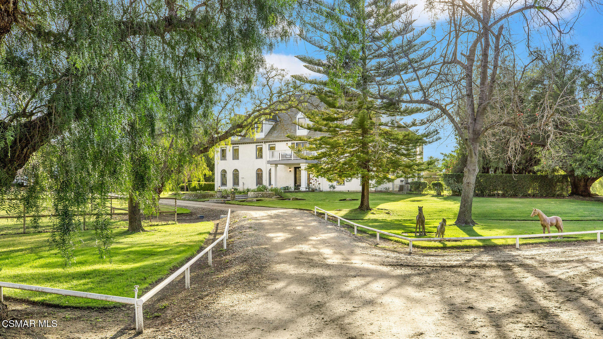 White two-story home beyond a tree-lined gravel drive, with a broad lawn and horse sculptures.
