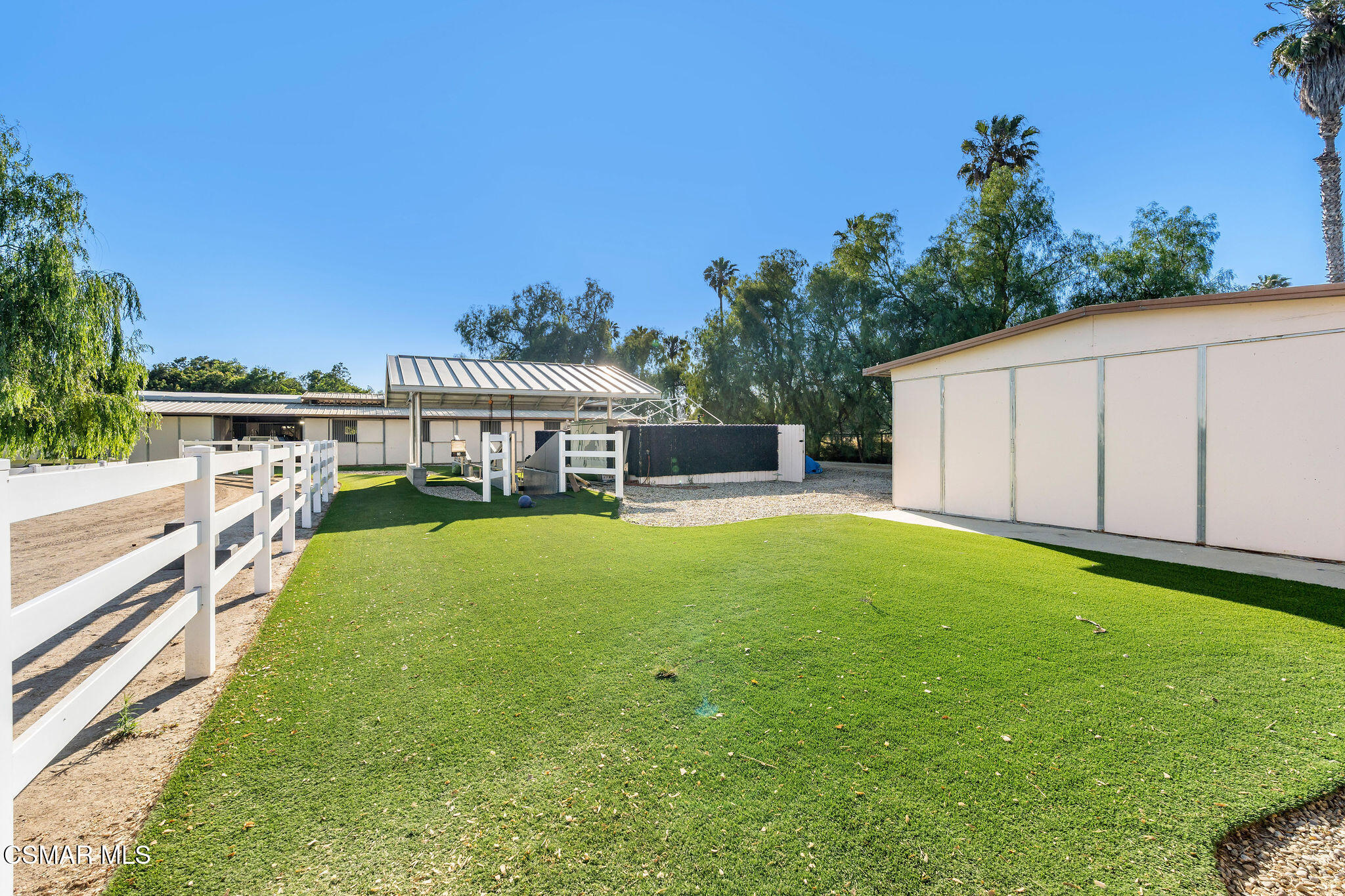 Green lawn bordered by white rail fencing, with a covered structure and detached outbuildings.