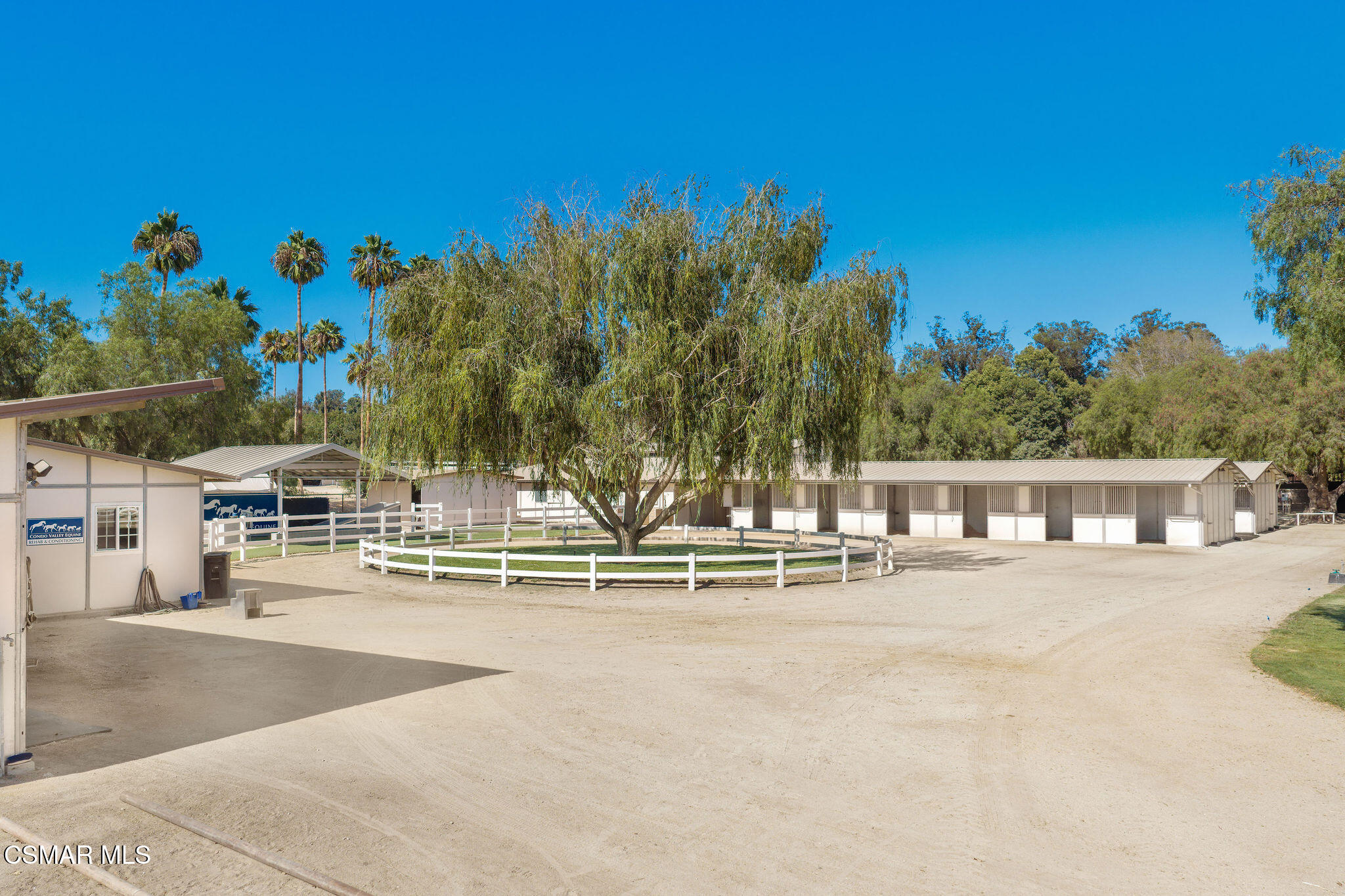 Equestrian facility with a long row of stalls and a fenced willow-tree enclosure.