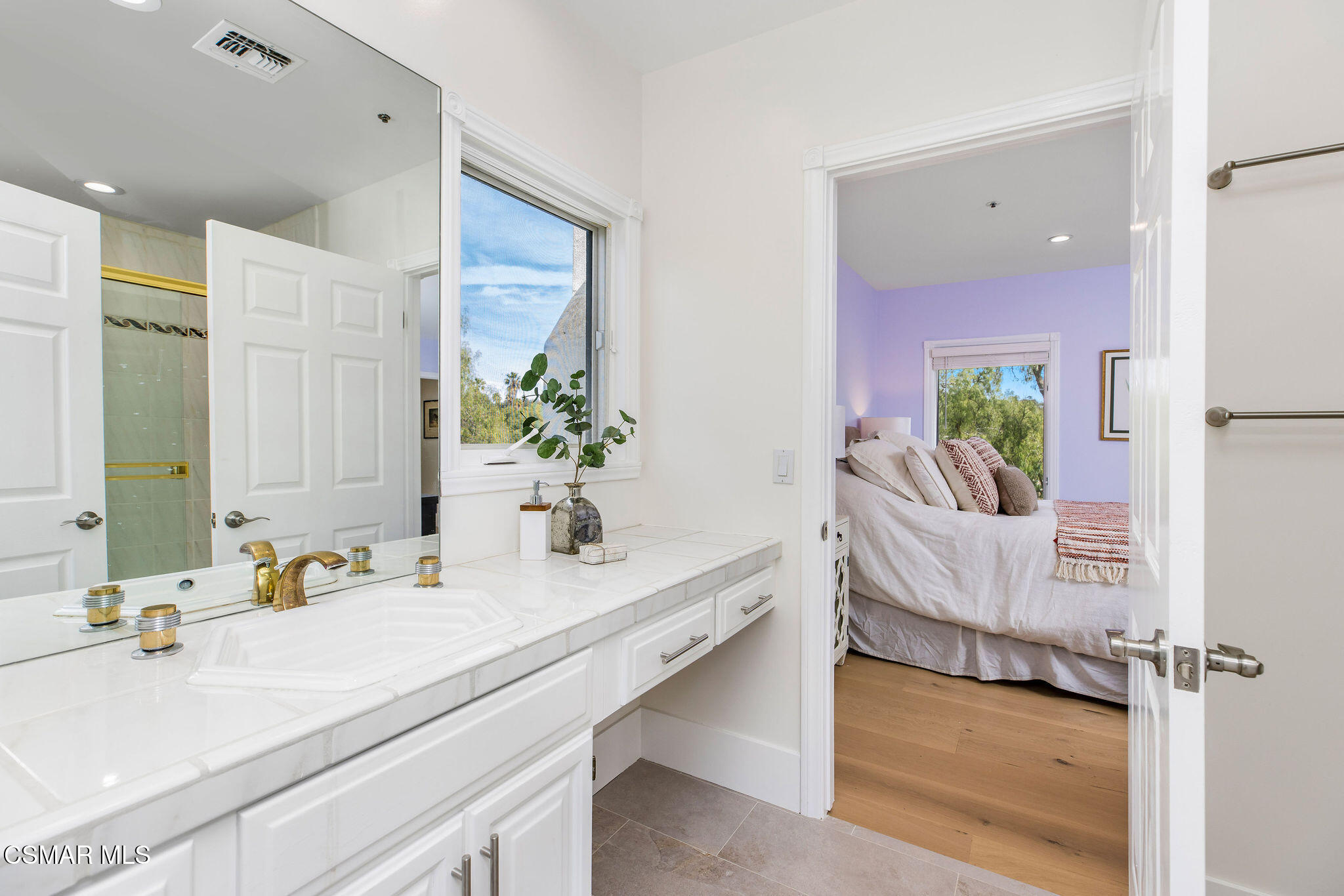 Bathroom vanity with white tile and gold-tone faucets, opening onto a lavender bedroom.