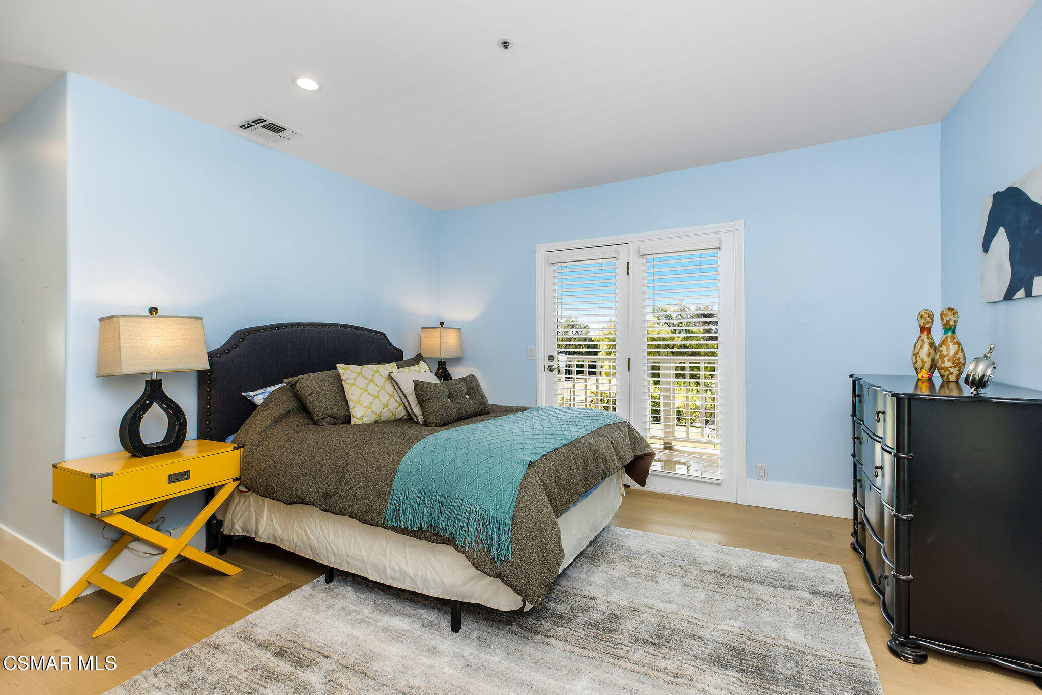 Bedroom with a dark upholstered bed, turquoise throw, pale blue walls, and white-shuttered glass doors.