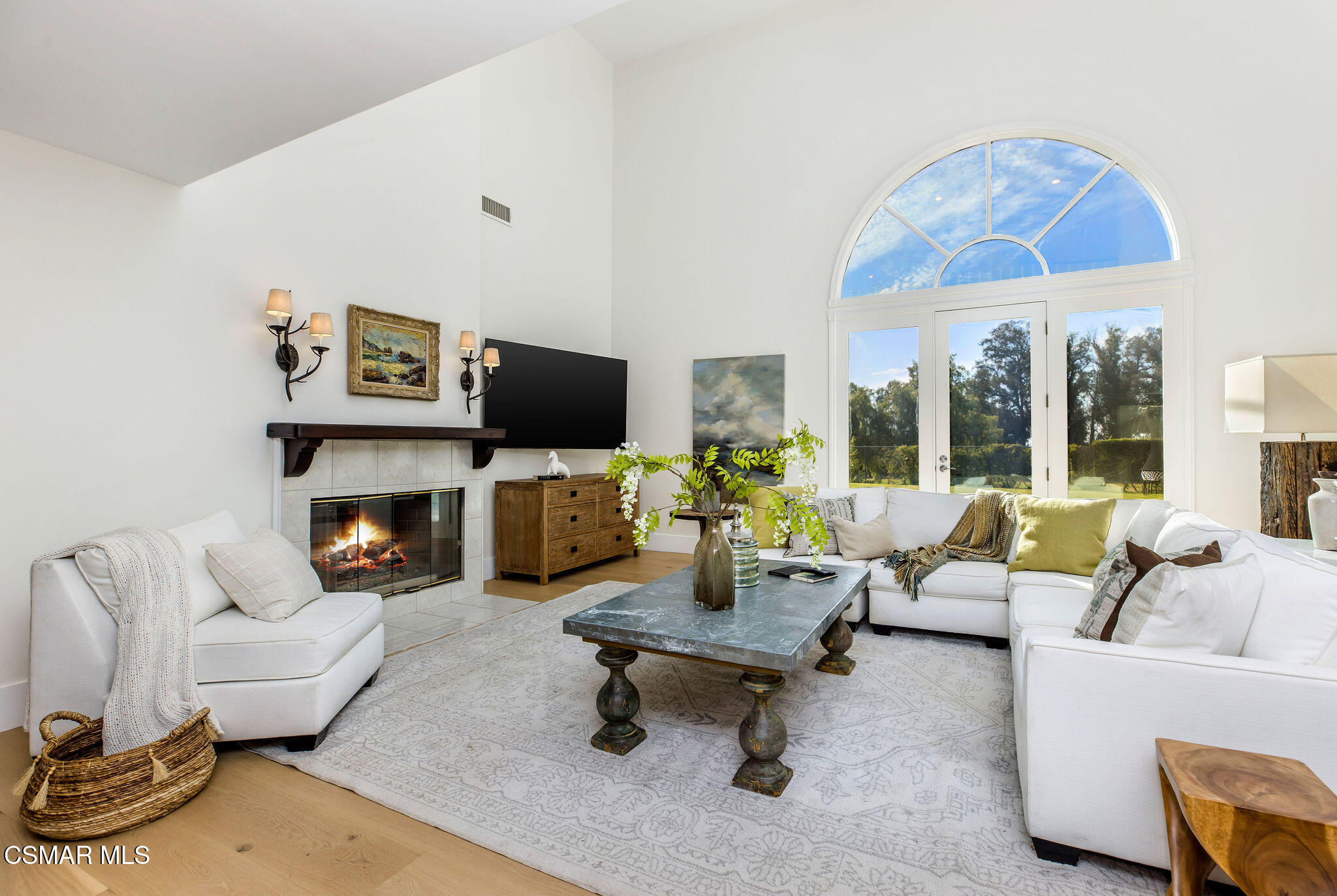 Living room with a lit fireplace, white seating, and an arched window above glass doors.