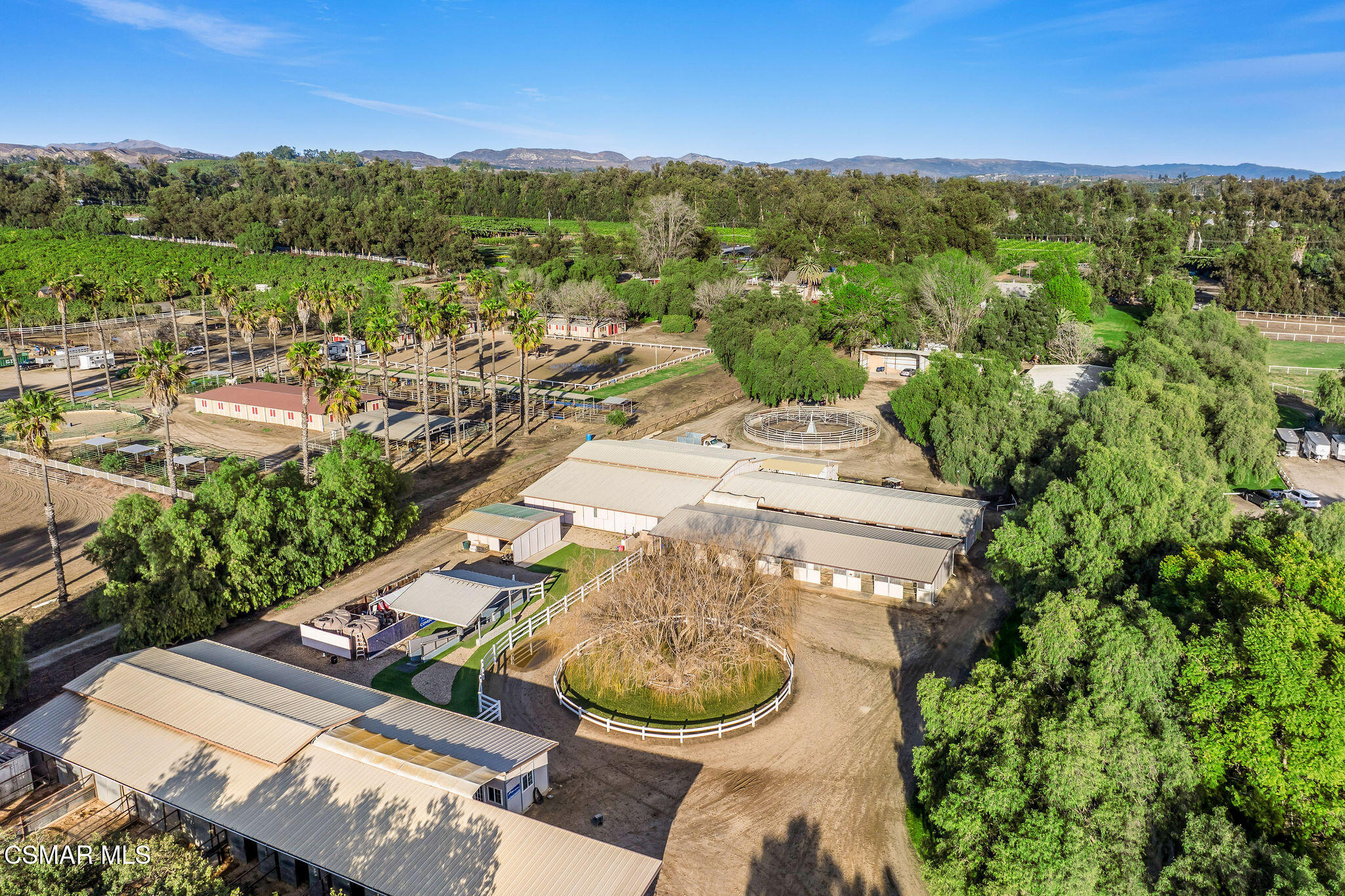 Aerial view of an equestrian property with barns, fenced paddocks, a circular horse walker, and distant mountains.