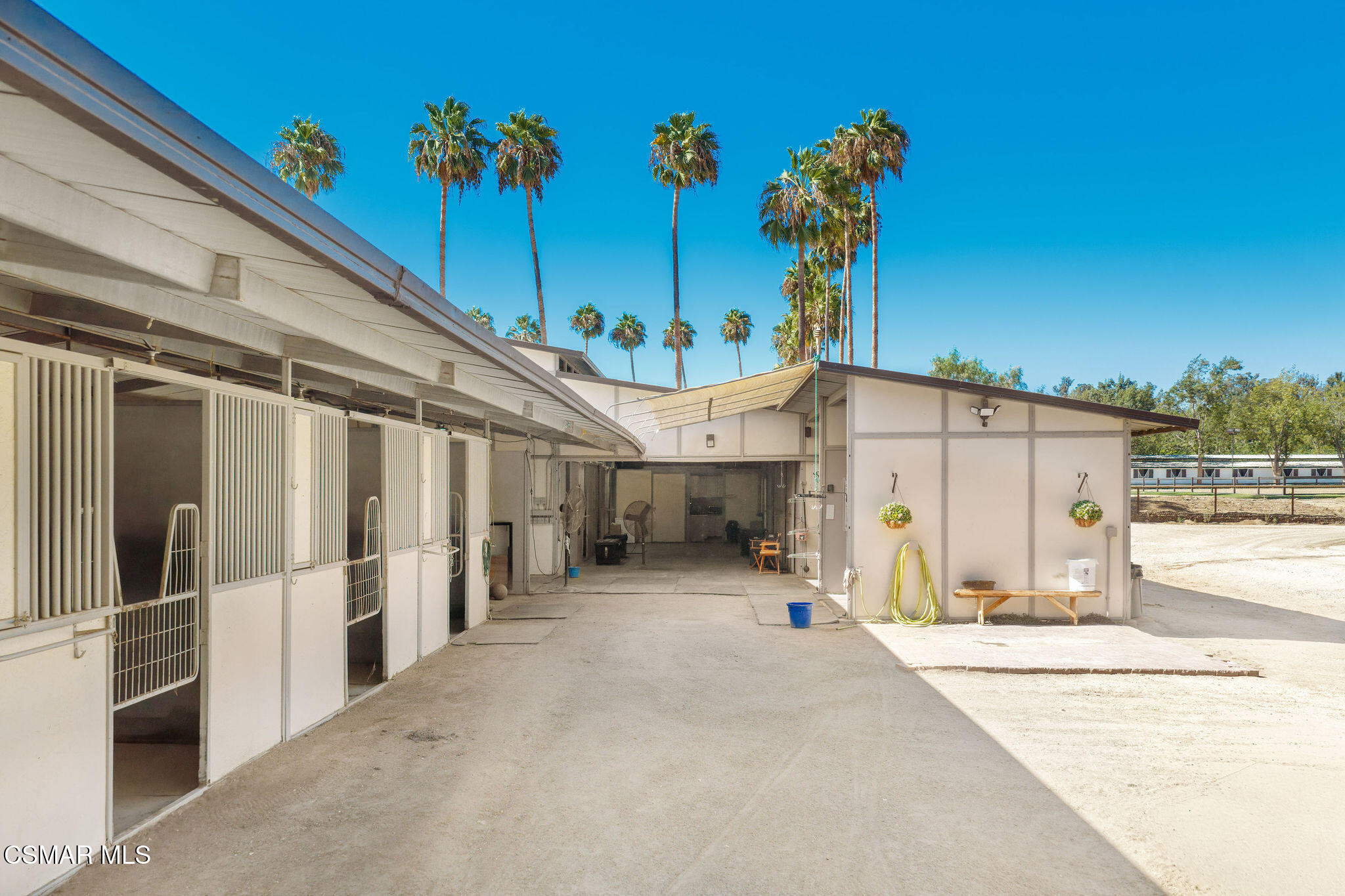 Stable courtyard with open horse stalls, a covered work area, and tall palm trees.
