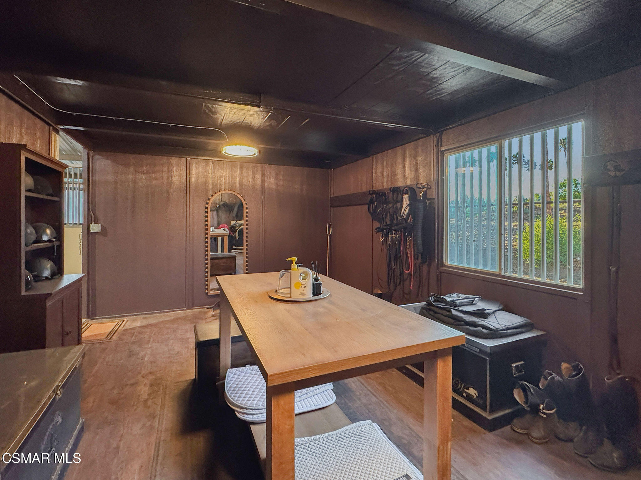 Tack room with a wooden table, hanging saddles and bridles, and helmets on shelves.