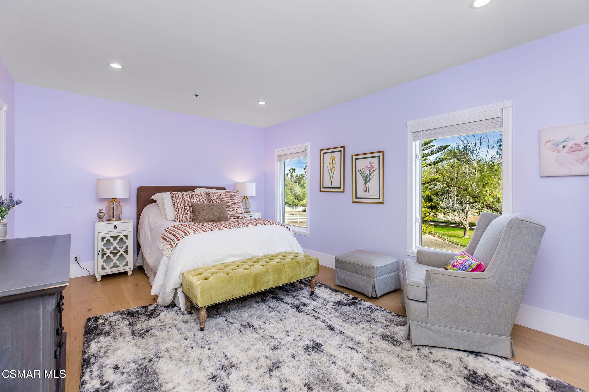 Bedroom with lavender walls, a white bed, green bench, and windows overlooking leafy grounds.