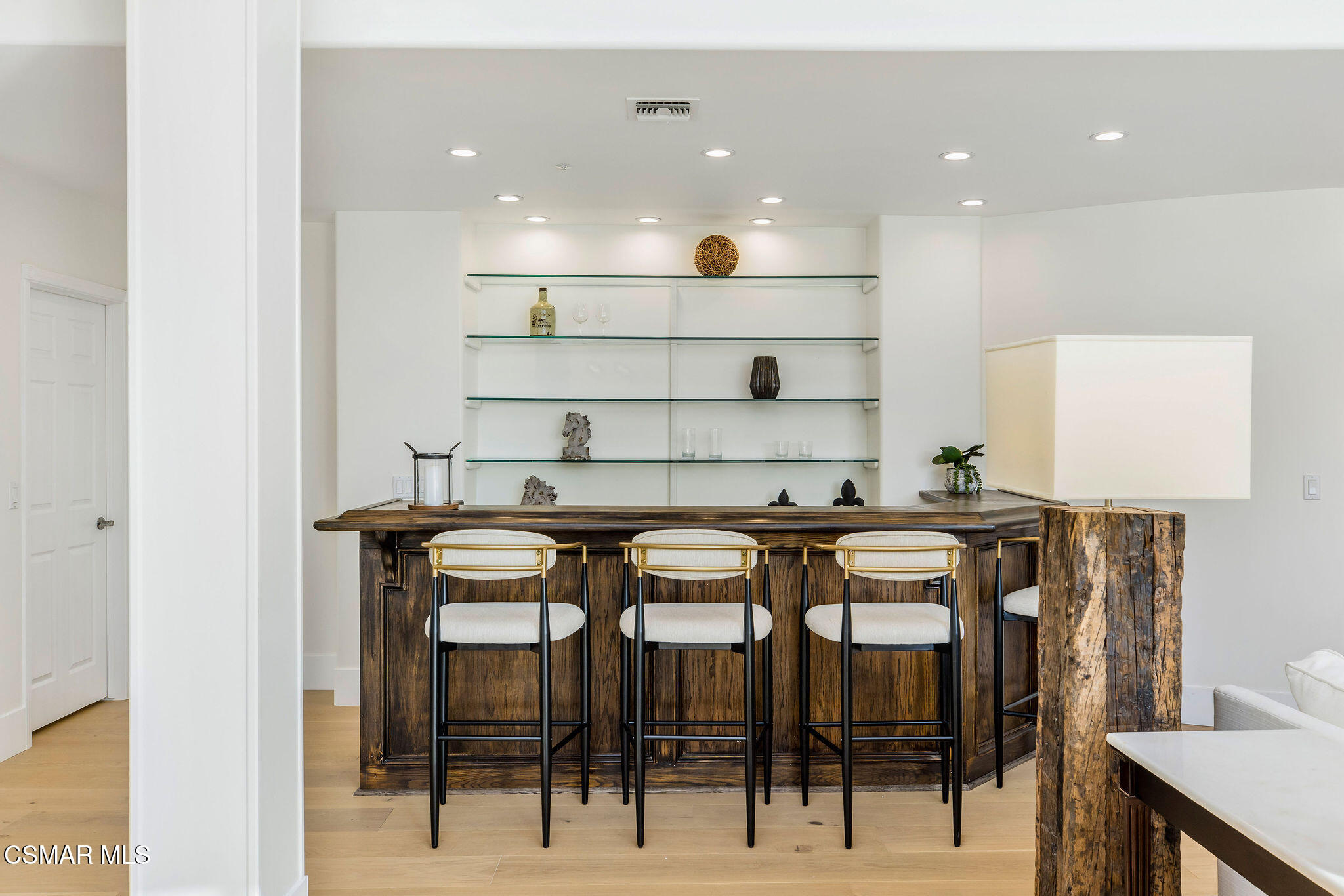 Interior bar with a rustic wood counter, three upholstered stools, and glass display shelves.