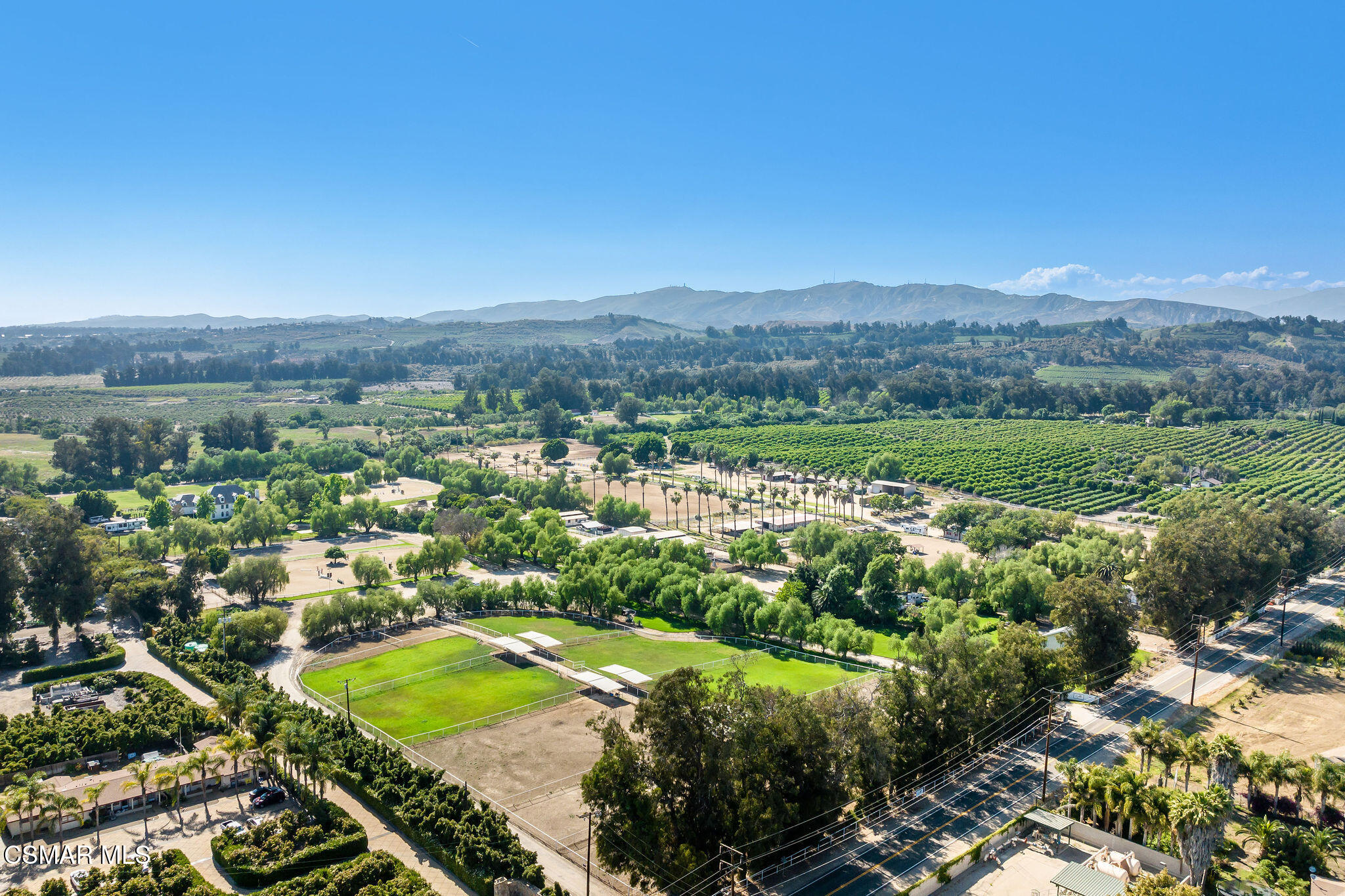 Aerial view of acreage with a fenced riding arena, vineyard rows, and distant mountains.