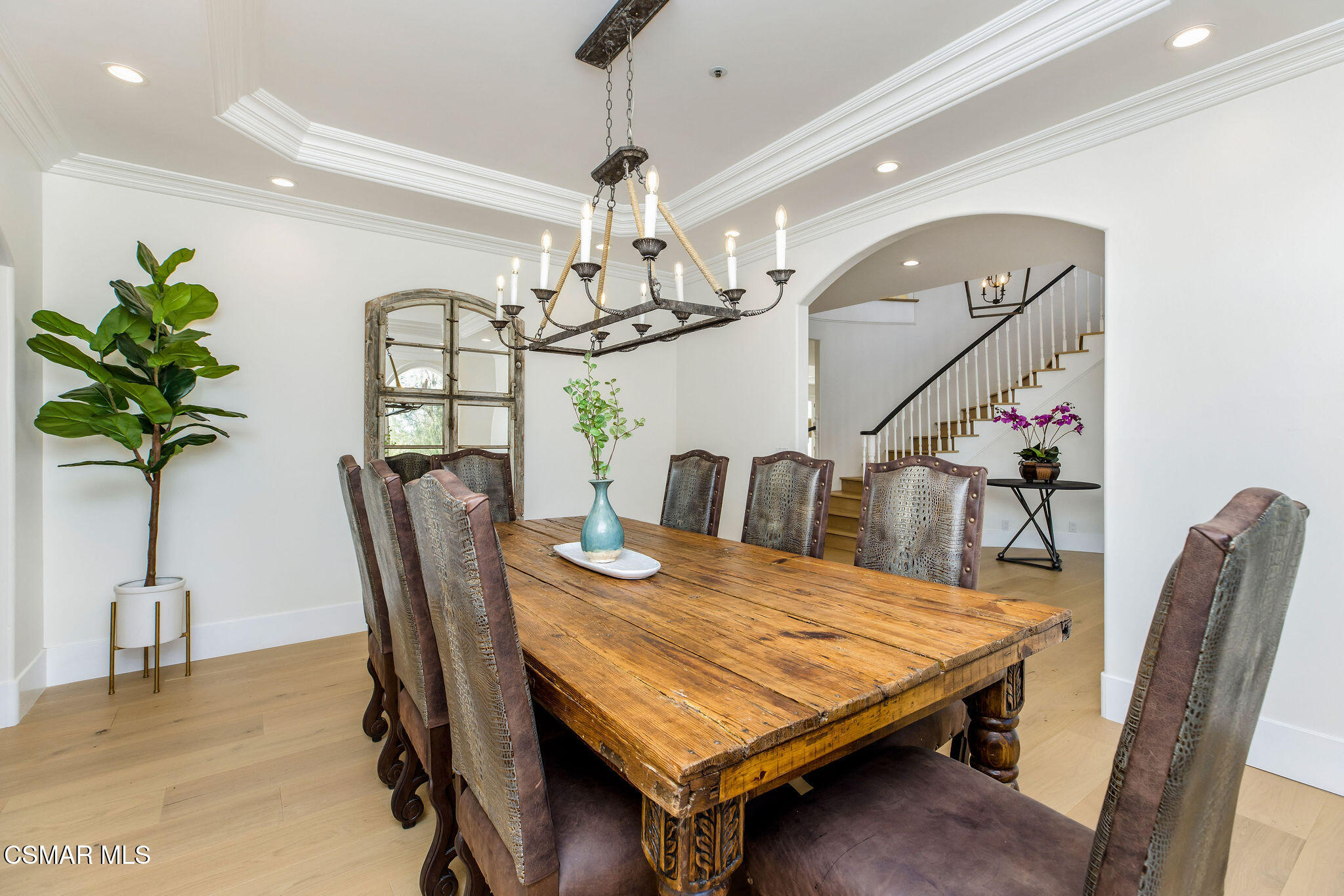 Dining room with a rustic wood table, upholstered chairs, and a chandelier beside an arched staircase opening.