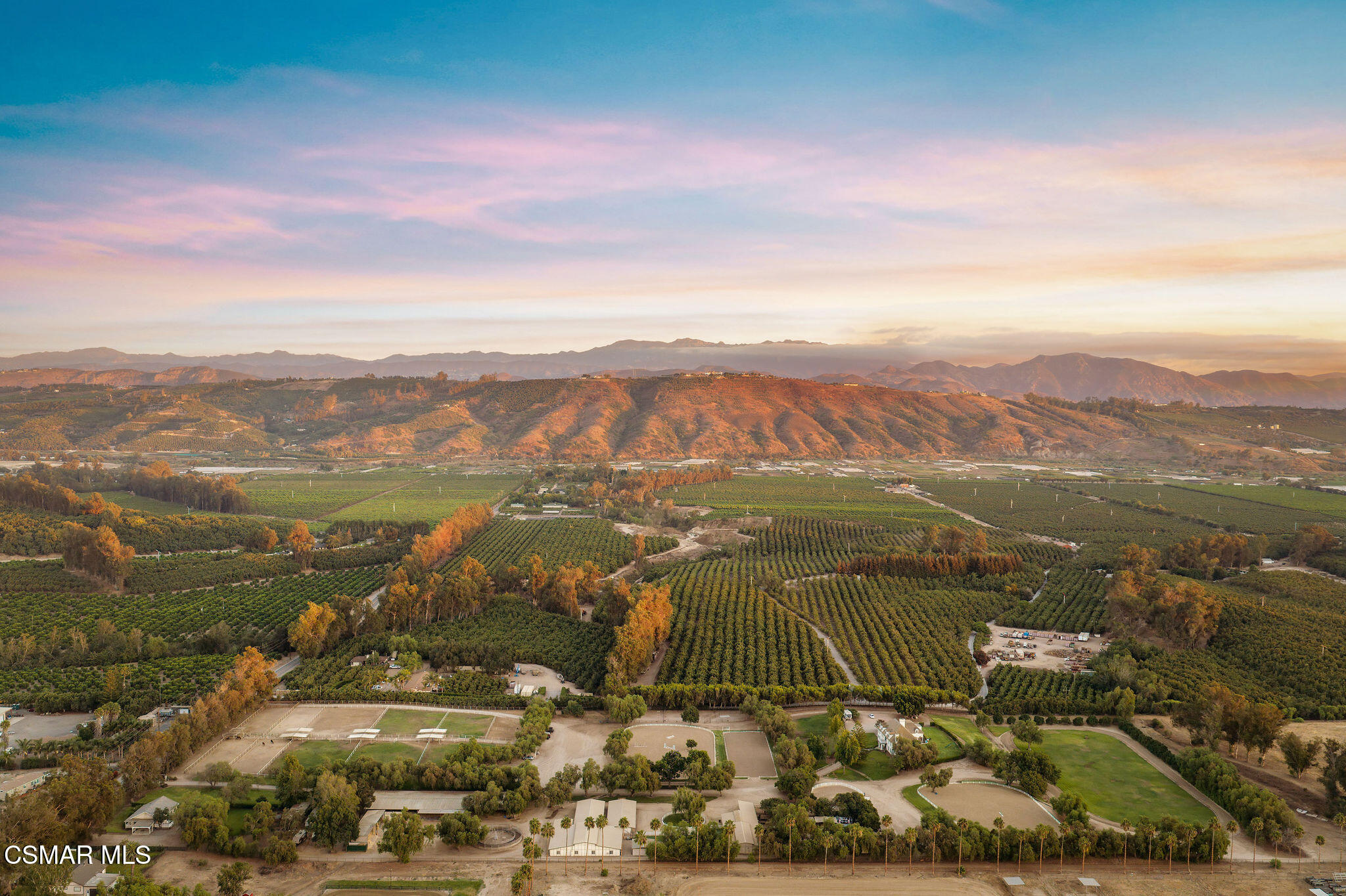 Aerial view of orchards and equestrian grounds beneath sunlit mountains.