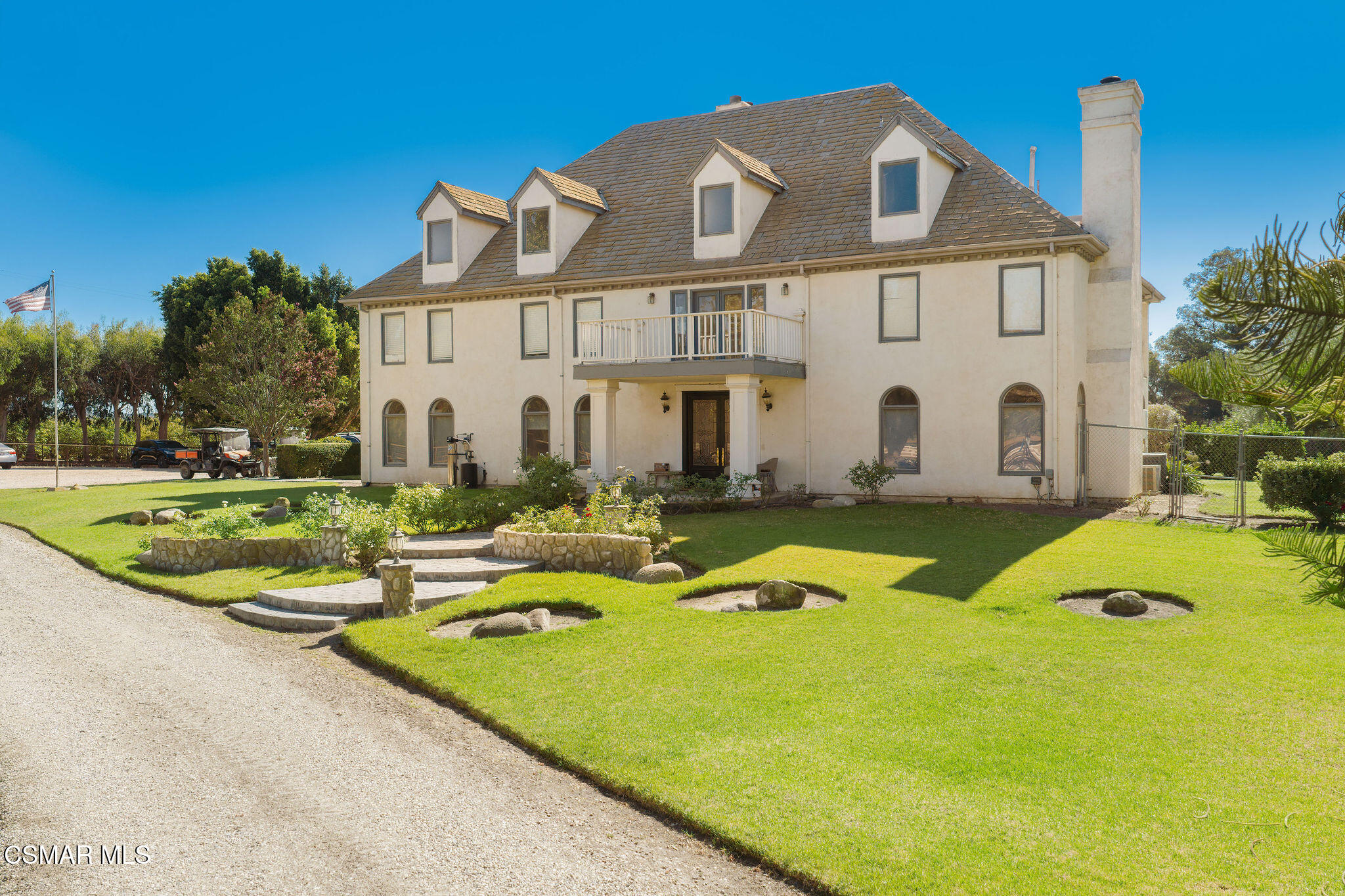 Large cream-colored house with roof dormers and balcony, surrounded by a landscaped lawn and gravel drive.