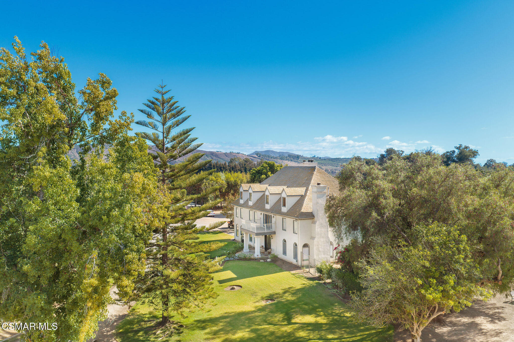 Aerial view of a white multi-story home amid mature trees, green lawns, and rolling hills.