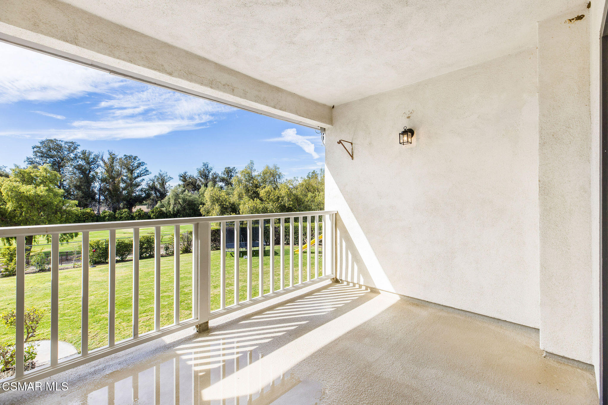 Covered balcony overlooking a green lawn and trees, with sunlight casting striped shadows across the floor.