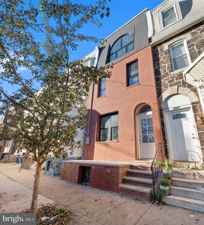 Red-brick Philadelphia rowhouse with an arched upper window and front steps beside a leafy sidewalk tree.