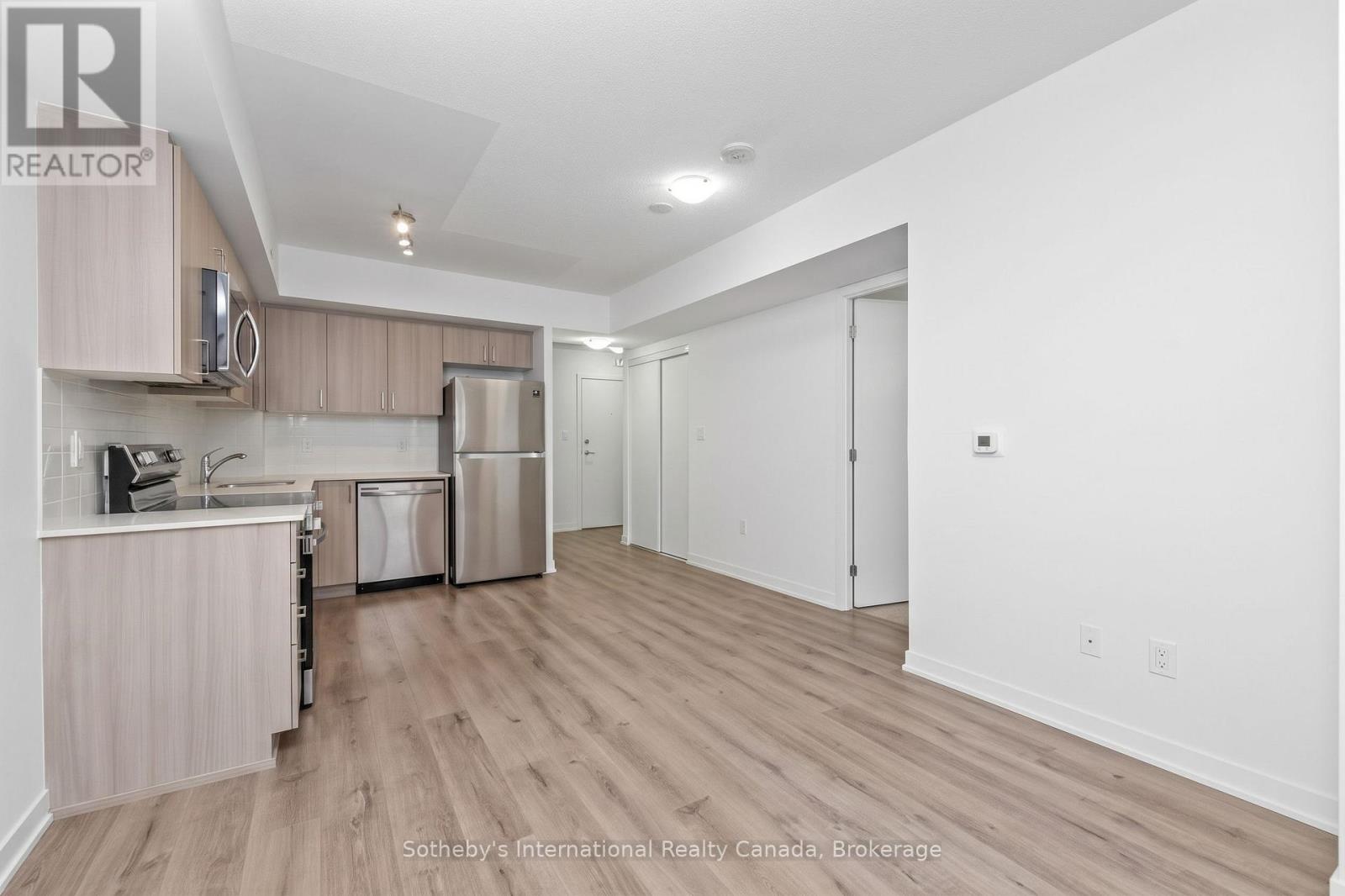 Unfurnished condo kitchen and living area with light wood cabinets, stainless steel appliances, and wood-look floors.