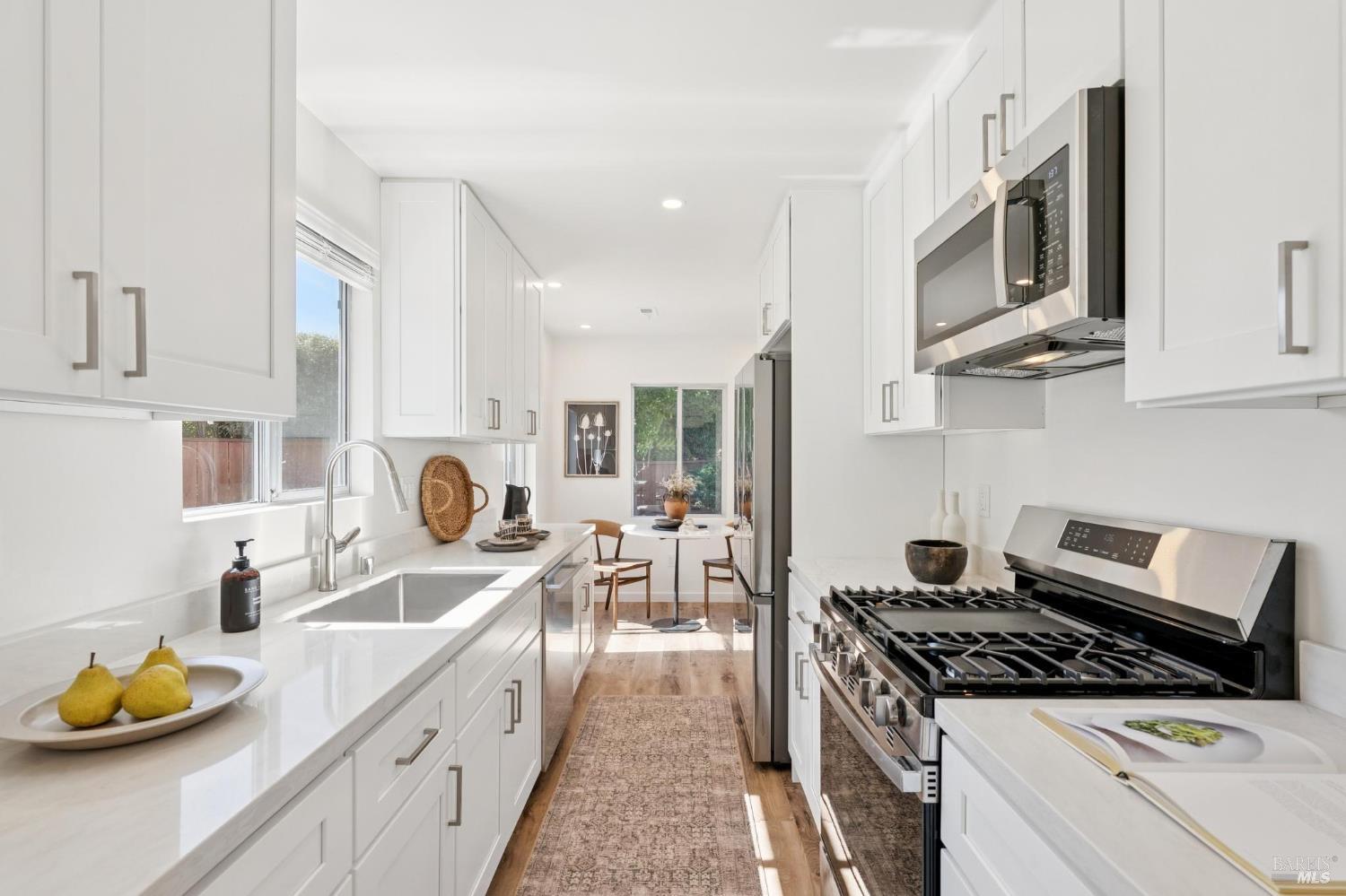 Bright white galley kitchen with a farmhouse sink, stainless steel range, and small dining area at the far end.