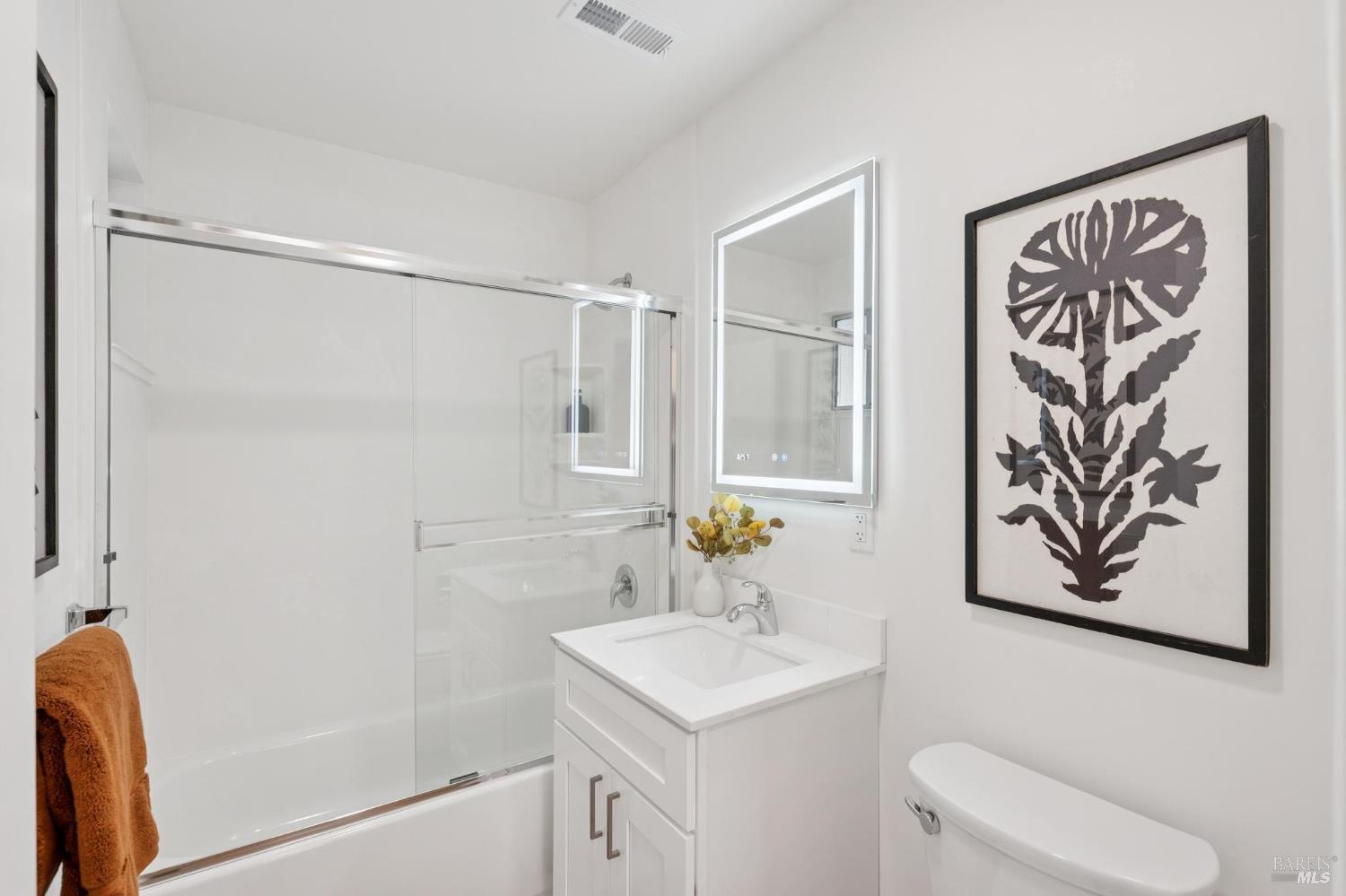 Bathroom with a glass-enclosed tub and shower, white vanity, illuminated mirror, and framed botanical print.