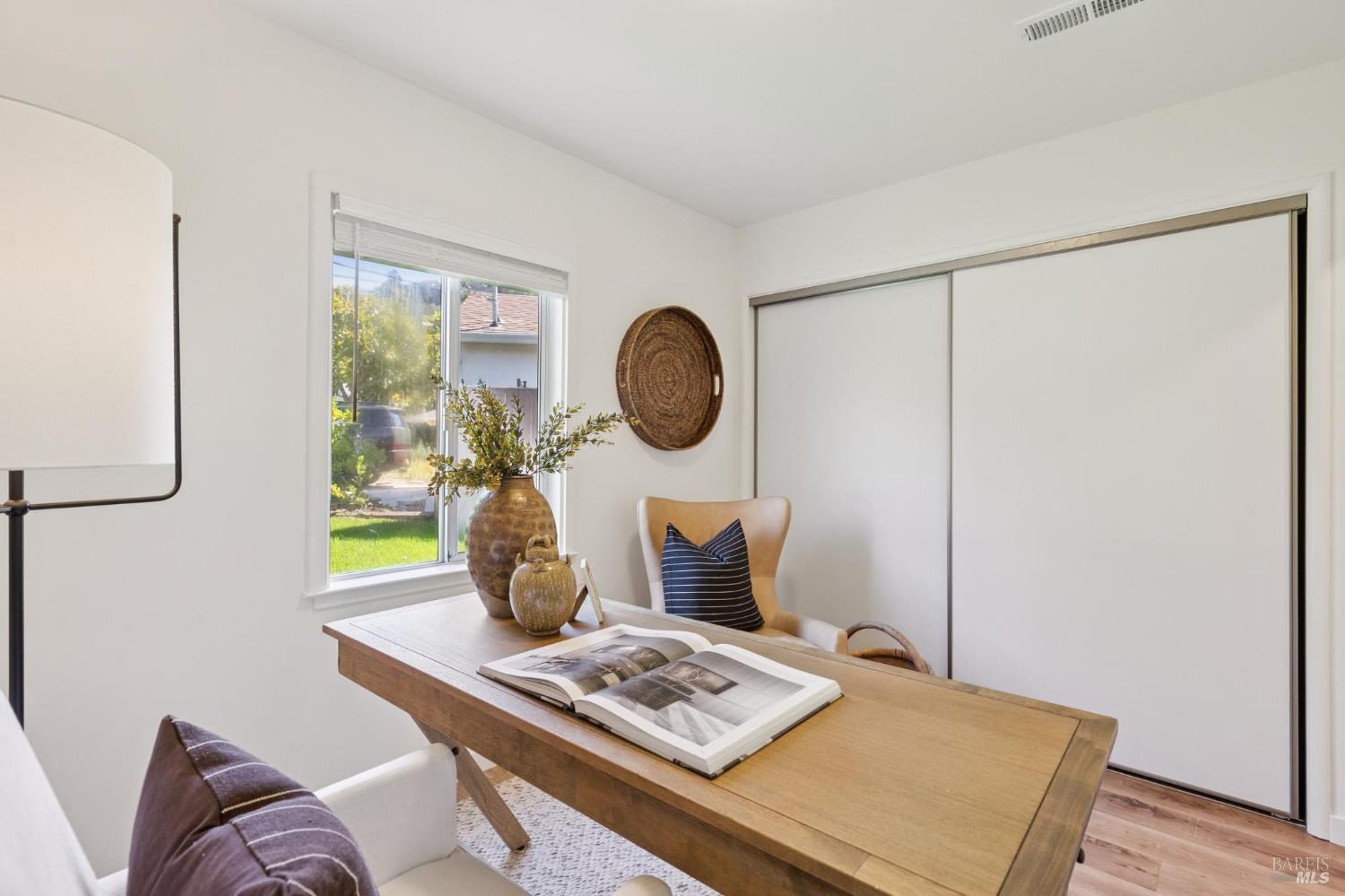 Home office with a wooden desk, open book, cushioned chair, and window overlooking a green yard.