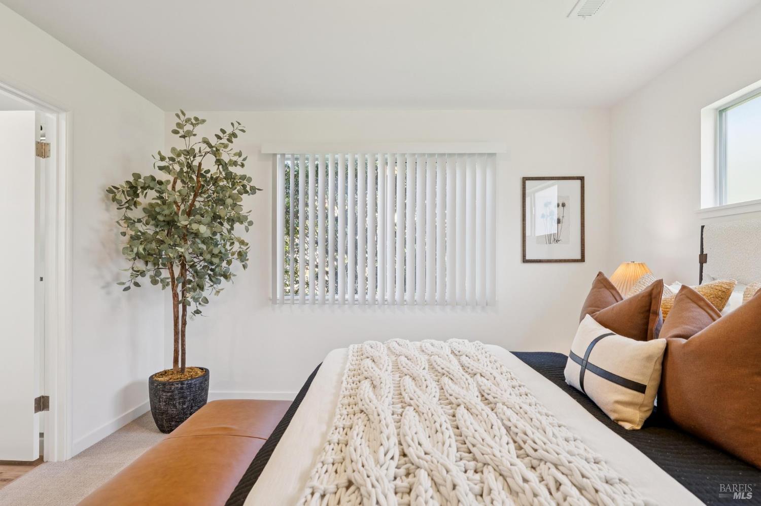 Bright bedroom with a chunky white knit blanket, brown pillows, and a potted tree.