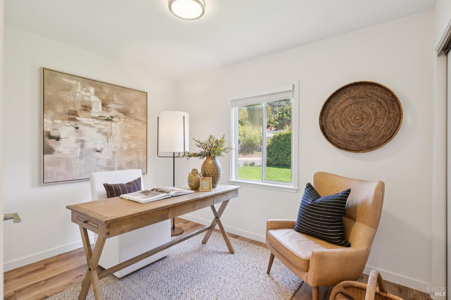 Staged home office with a wooden desk, tan armchair, and window overlooking greenery.