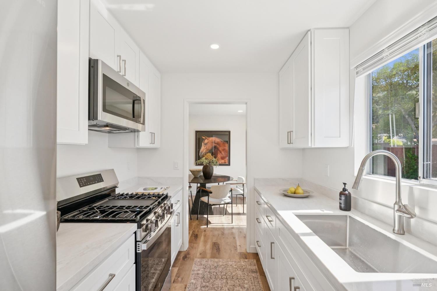 White galley kitchen with stainless steel range and sink beneath a window, opening to a dining area.