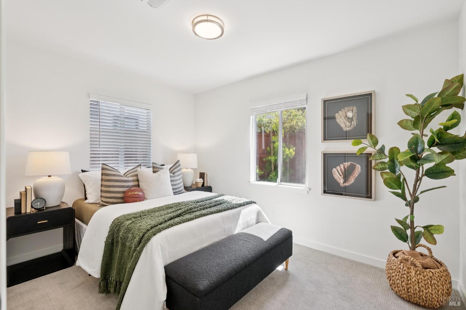 Staged bedroom with a white bed, green throw, baseball-themed artwork, and a large potted plant.