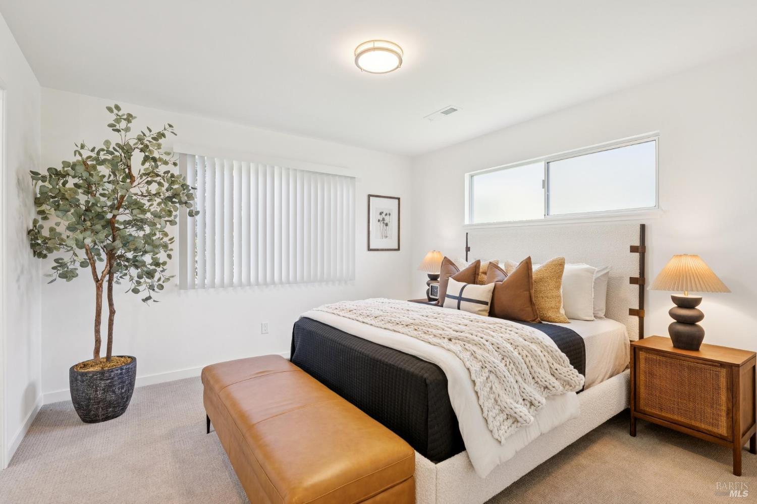 Staged bedroom with a king-size bed, leather bench, potted tree, and wide windows.