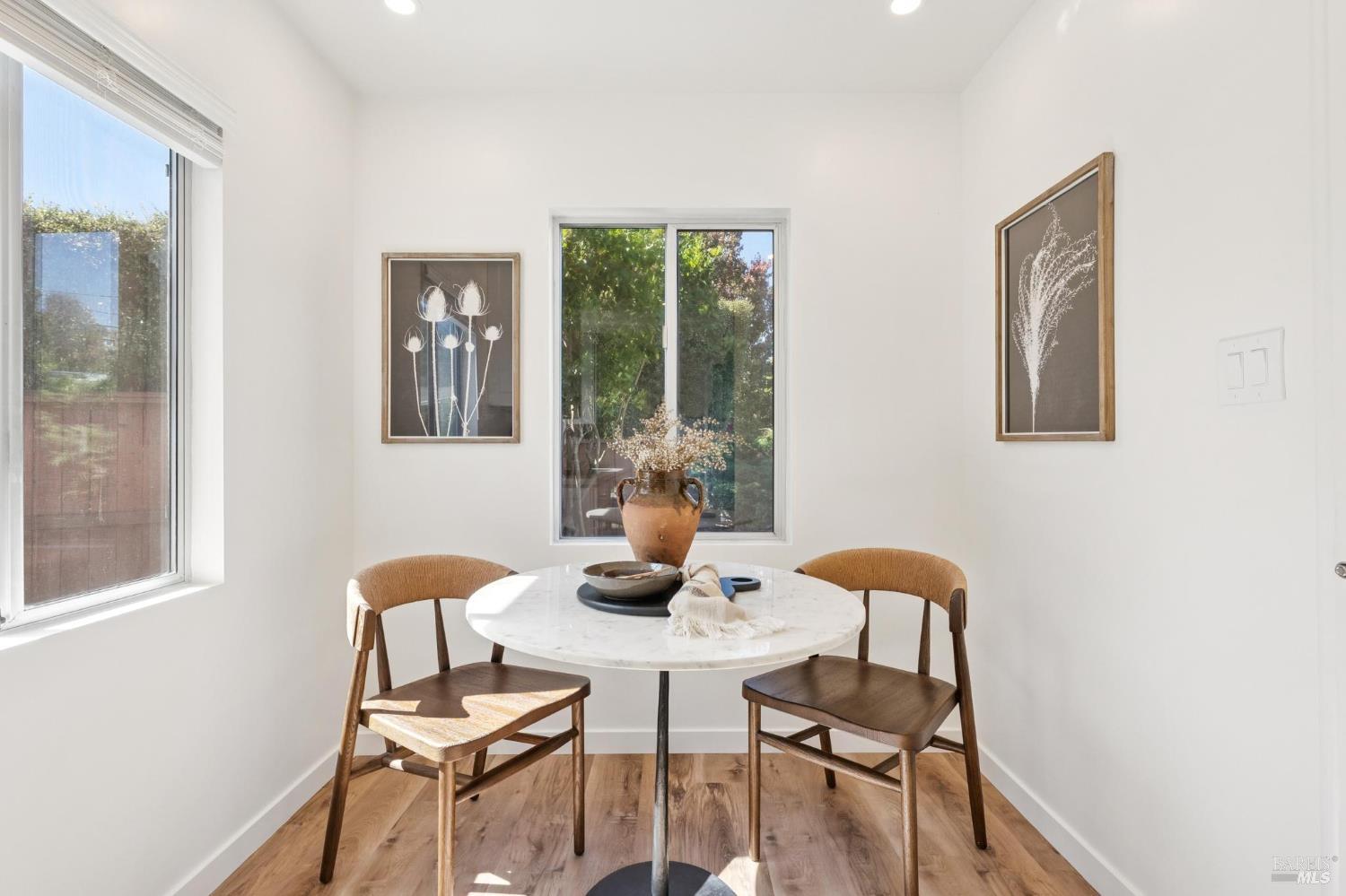 Bright dining nook with a round white table, two wooden chairs, and windows overlooking greenery.