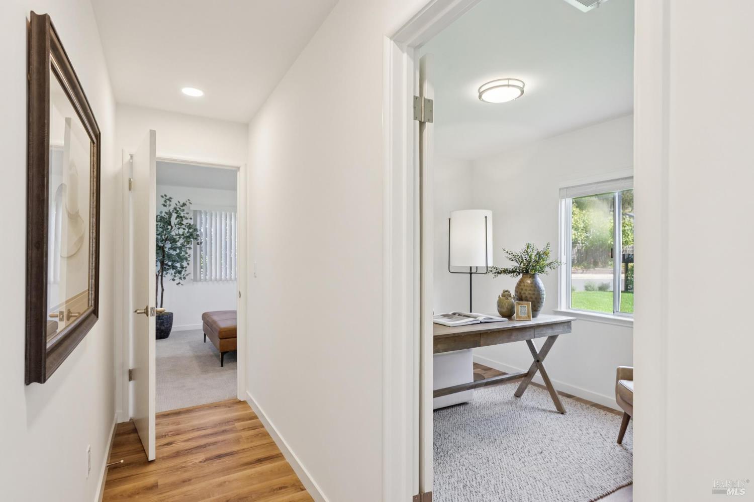 Hallway opening to a furnished home office with a desk and window, beside a carpeted sitting room.