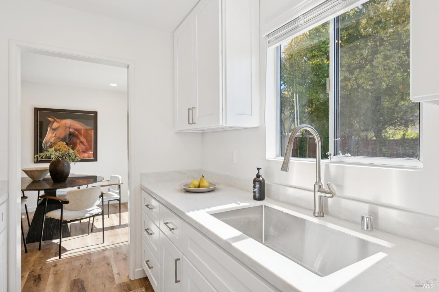 White kitchen with a large sink beneath a window and an adjoining dining area.