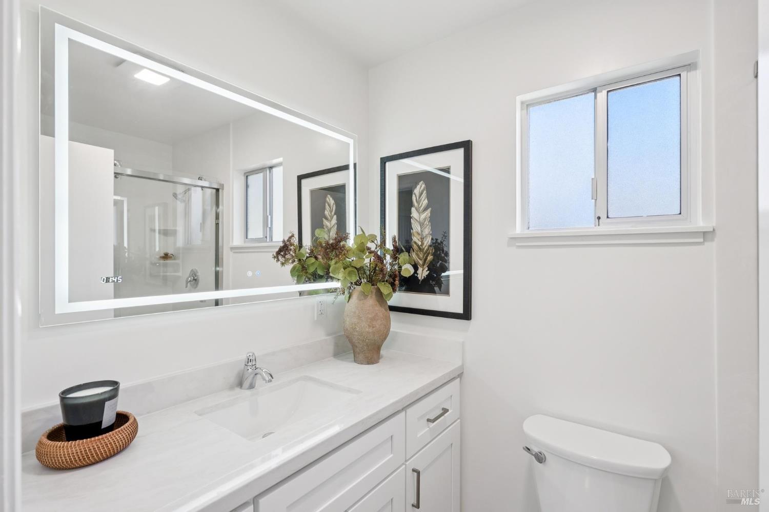 Bright bathroom with a long white vanity, illuminated mirror, glass shower, and window.
