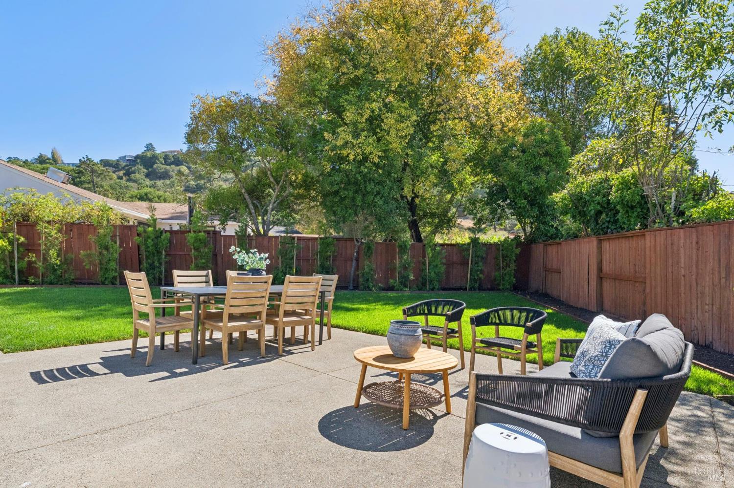 Backyard patio with dining and lounge seating beside a lawn and mature trees.