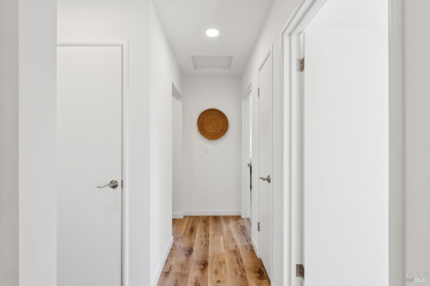 Bright white hallway with wood-look floors, several doors, and a round woven wall decoration.