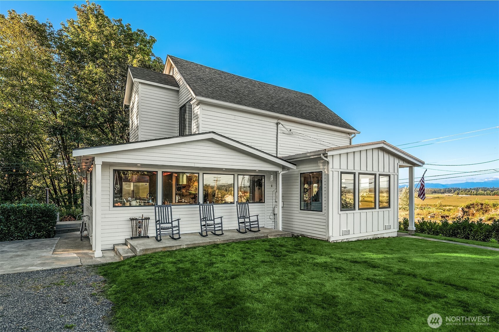 White two-story house with a covered porch and rocking chairs, overlooking open fields and distant mountains.