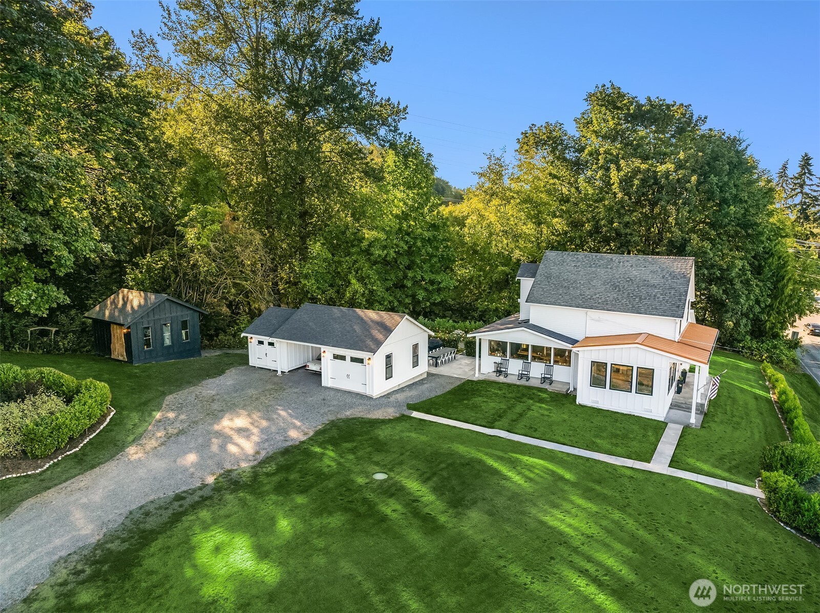 Aerial view of a white farmhouse, detached garage, and shed on a broad lawn among mature trees.