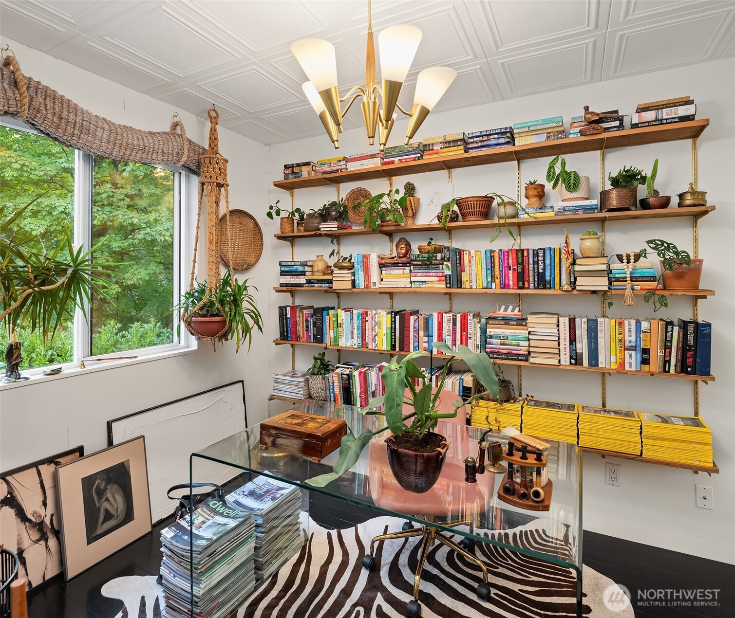 Home office with book-filled wooden shelves, a glass desk, hanging plants, and a large garden-facing window.