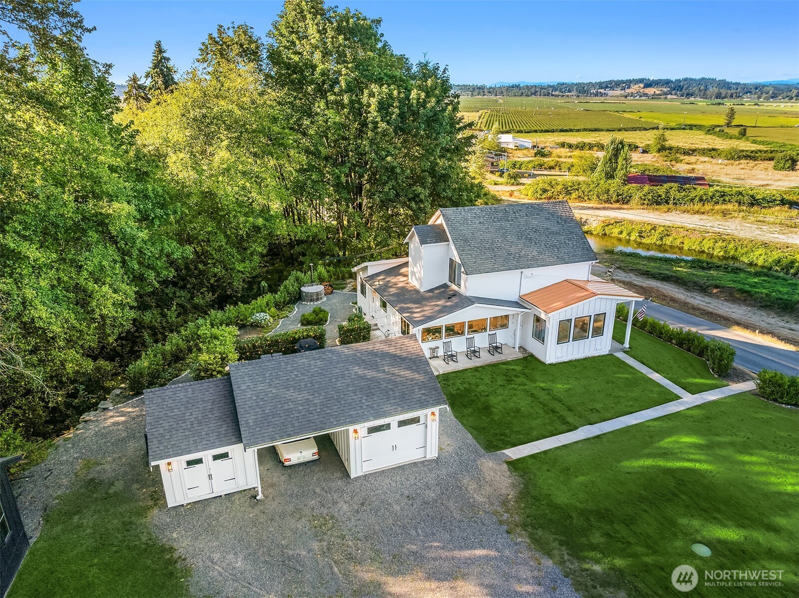 Aerial view of a white home and detached garage bordered by trees, lawn, and open farmland.