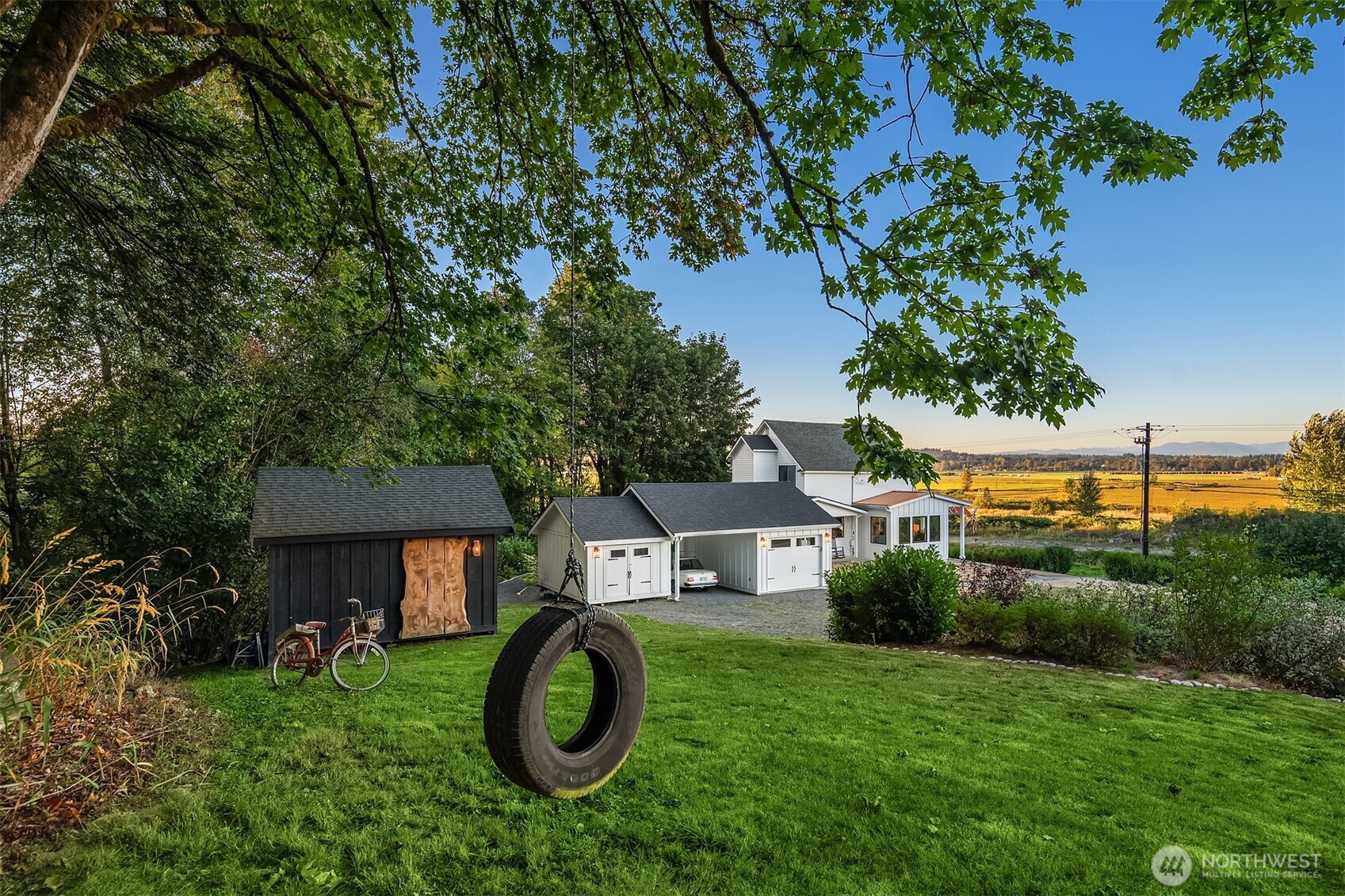 White house and detached garage beside a green lawn, tire swing, shaded shed, and open fields.