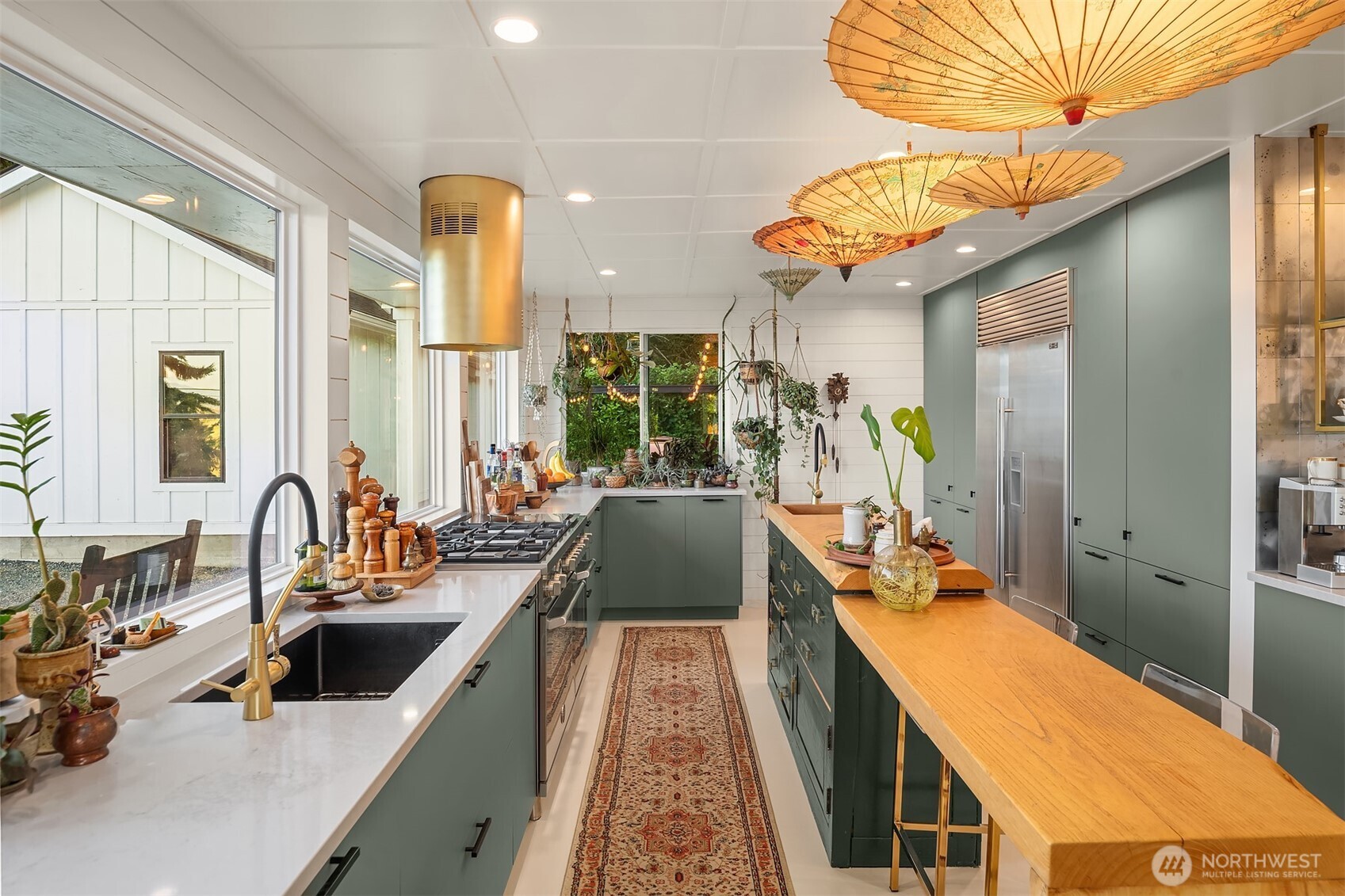 Long kitchen with green cabinets, a wood-topped island, hanging paper parasols, and a patterned runner.