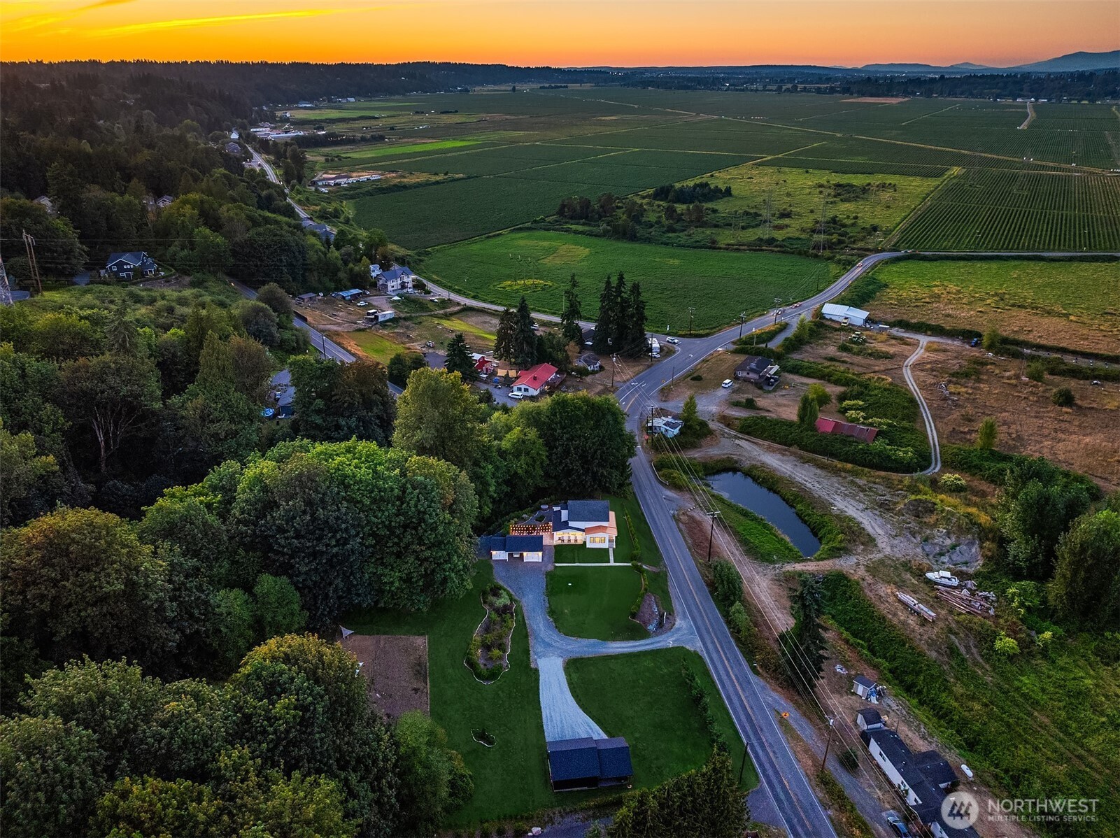 Rural home and outbuildings beside a road, surrounded by farmland beneath a colorful sunset sky.