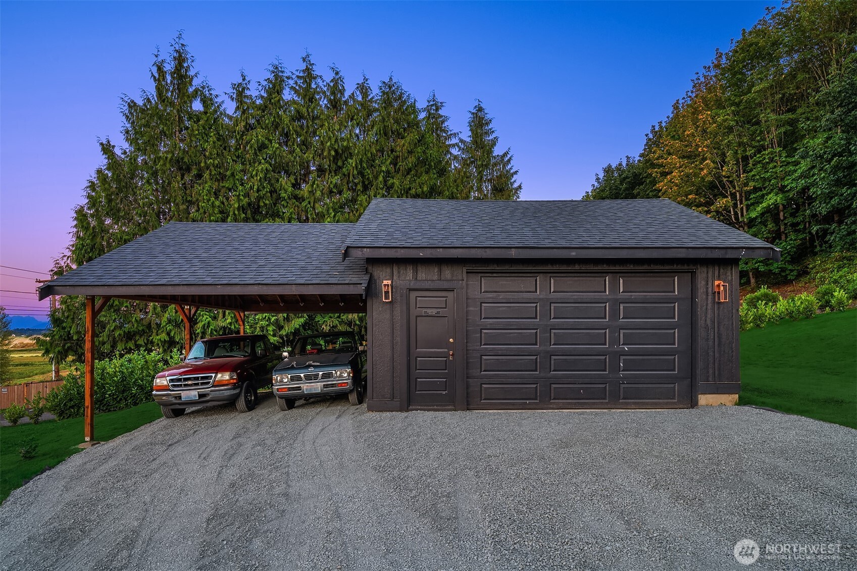 Dark detached garage with a two-car carport, gravel driveway, and wooded surroundings at dusk.